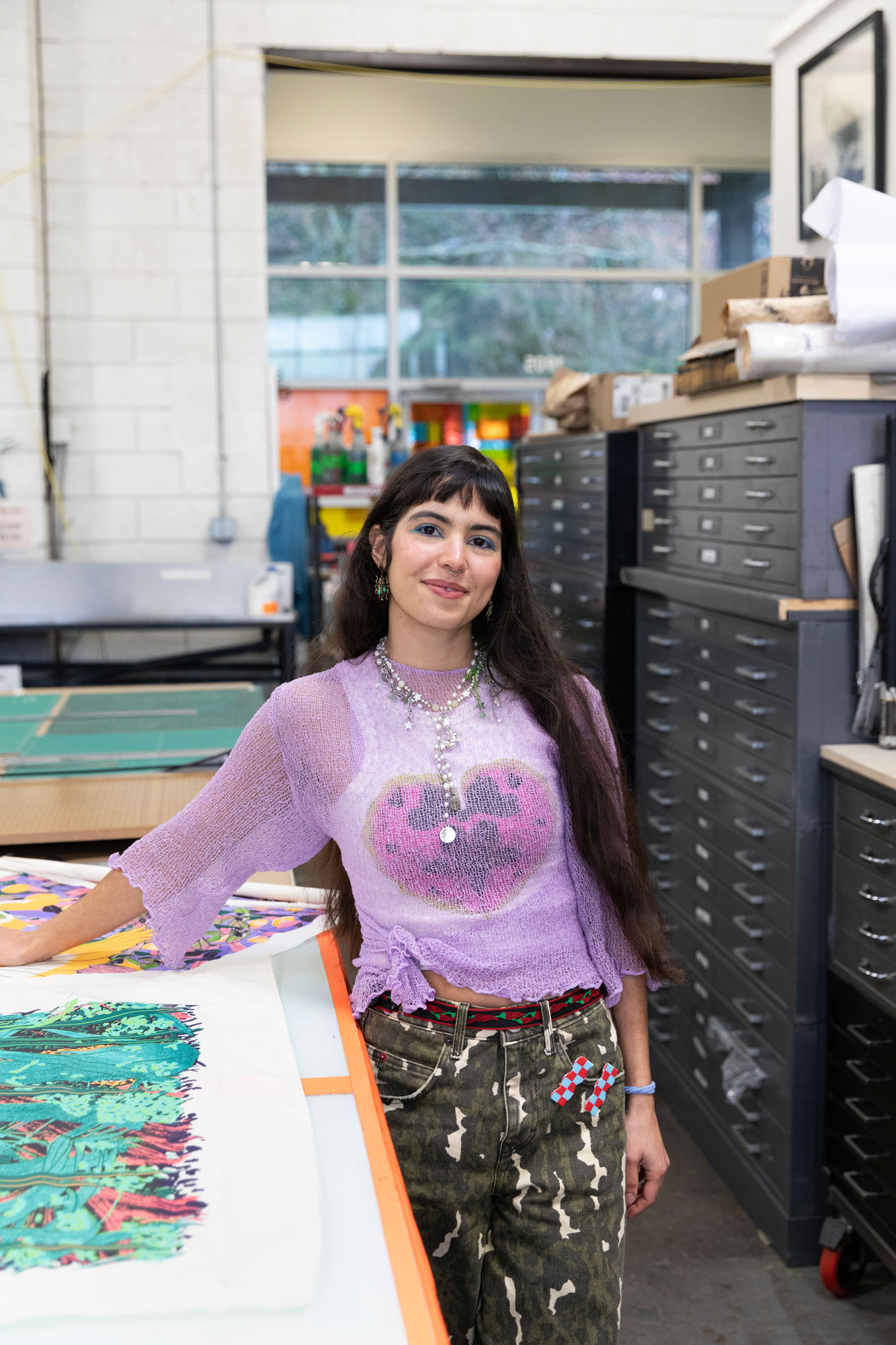 Artist María Zamora standing by a table with her screenprints on it and tall flat storage drawer cabinets behind her