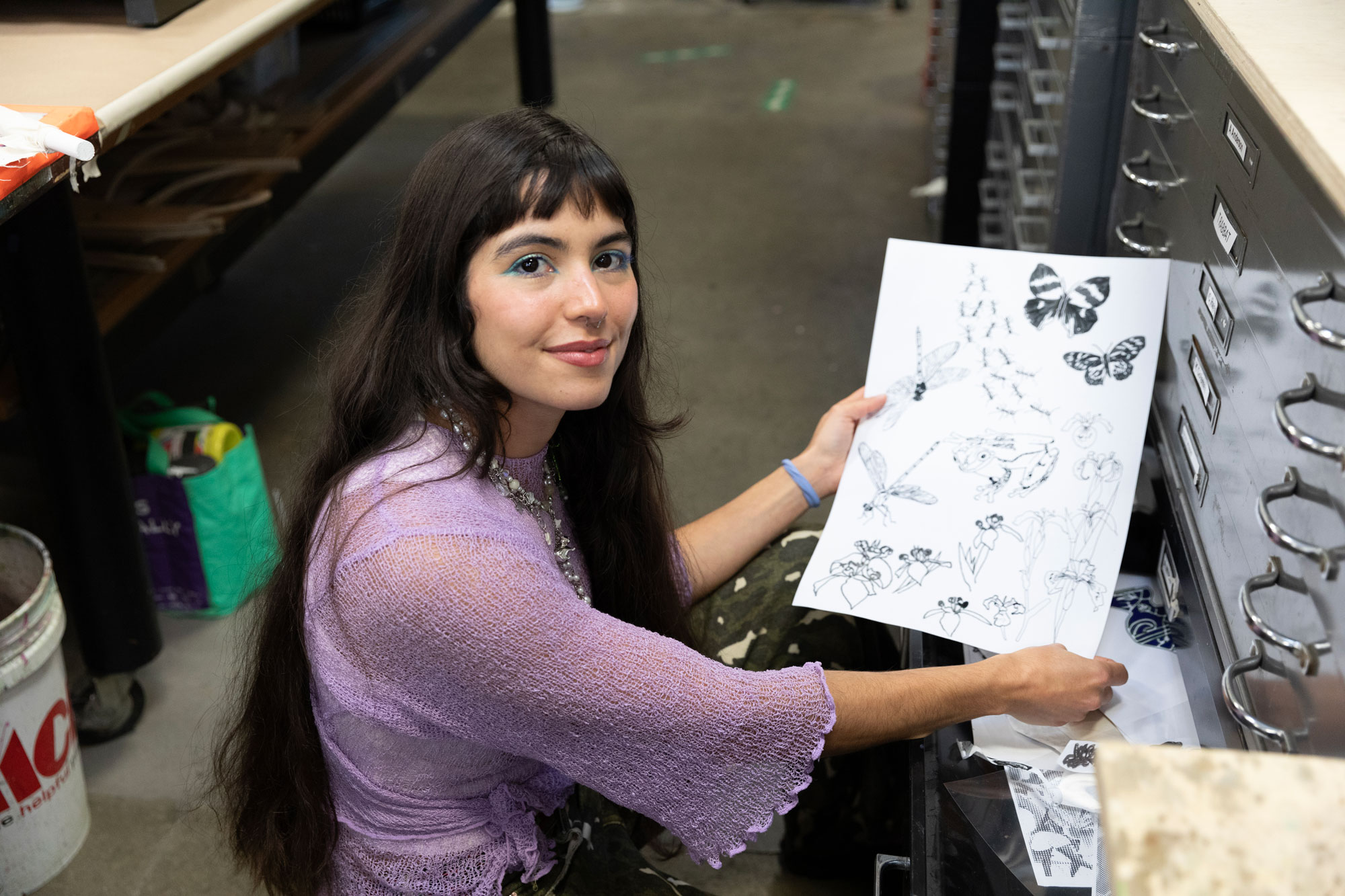 Artist María Zamora squatting by an open drawer showing a sheet with drawings of flora and fauna
