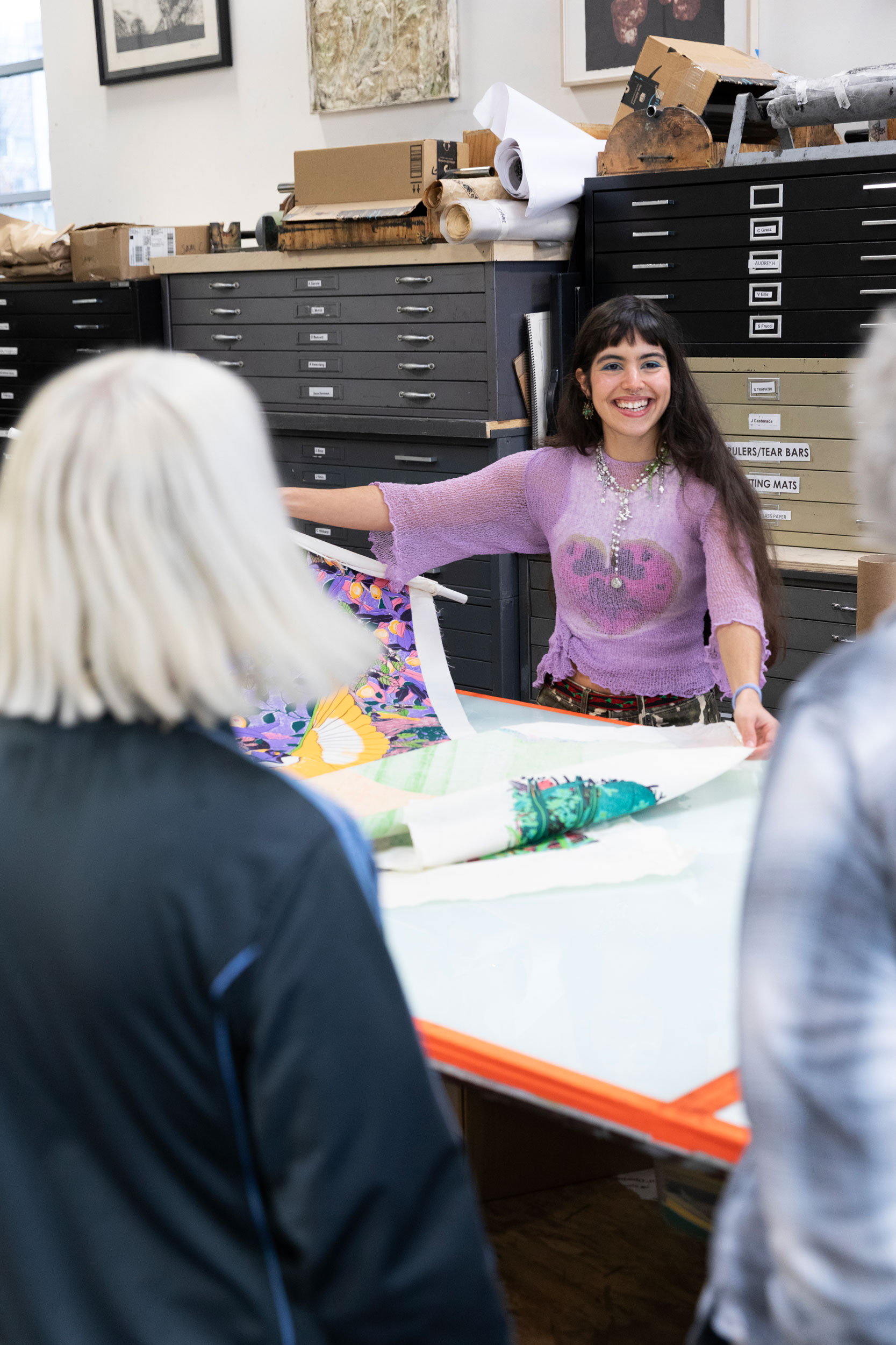 Artist María Zamora smiling while showing people some of her brightly colored screenprints on a table