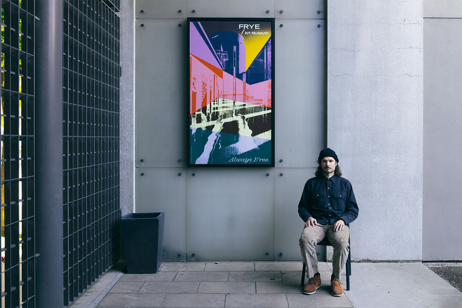A man in a blue shift and grey pants sitting next to a large poster on a wall in an outdoor courtyard