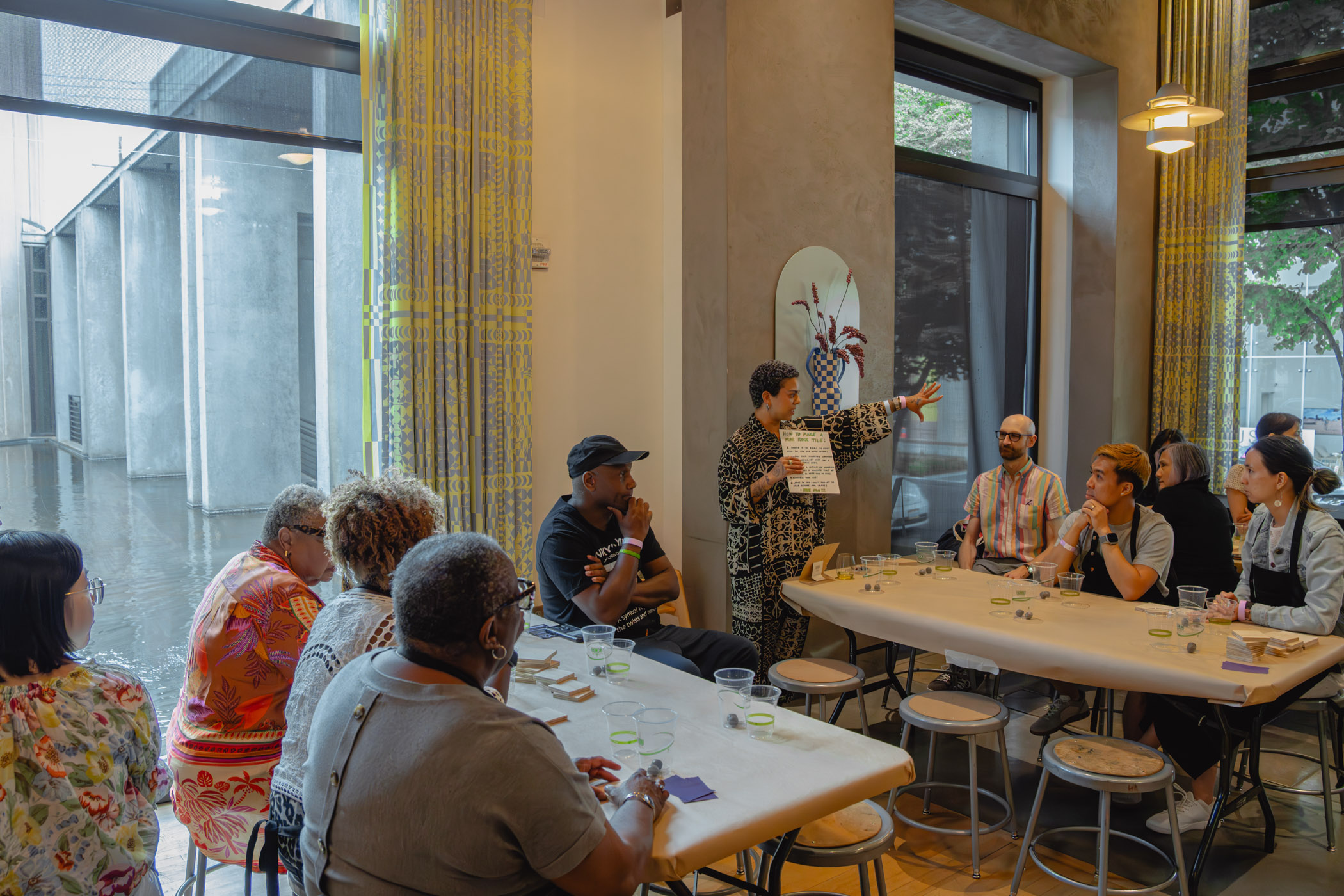 A large group of people seated at different tables listening to a person explain how to to an art activity