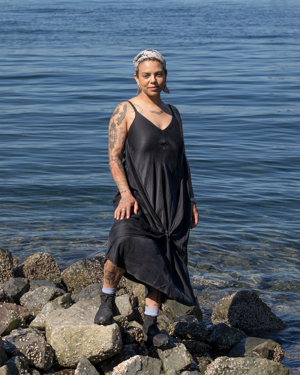 Emma Kates-Shaw standing on a rocky beach with the water behind them and looking directly at the camera