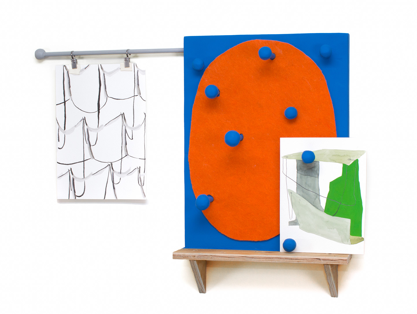 An artwork mounted on a white wall made of bits of household material painted primarily blue and orange. Pieces of paper with different designs are attached.