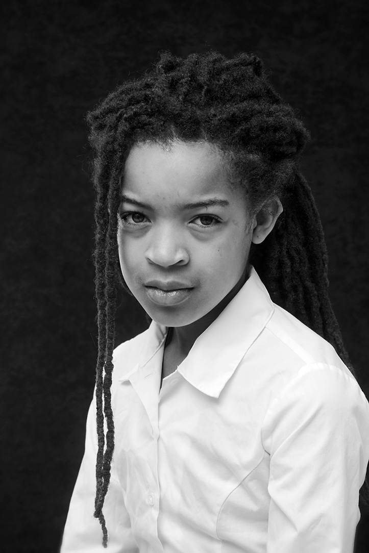A formal portrait style photograph of a child with long hair in a white, button up shirt