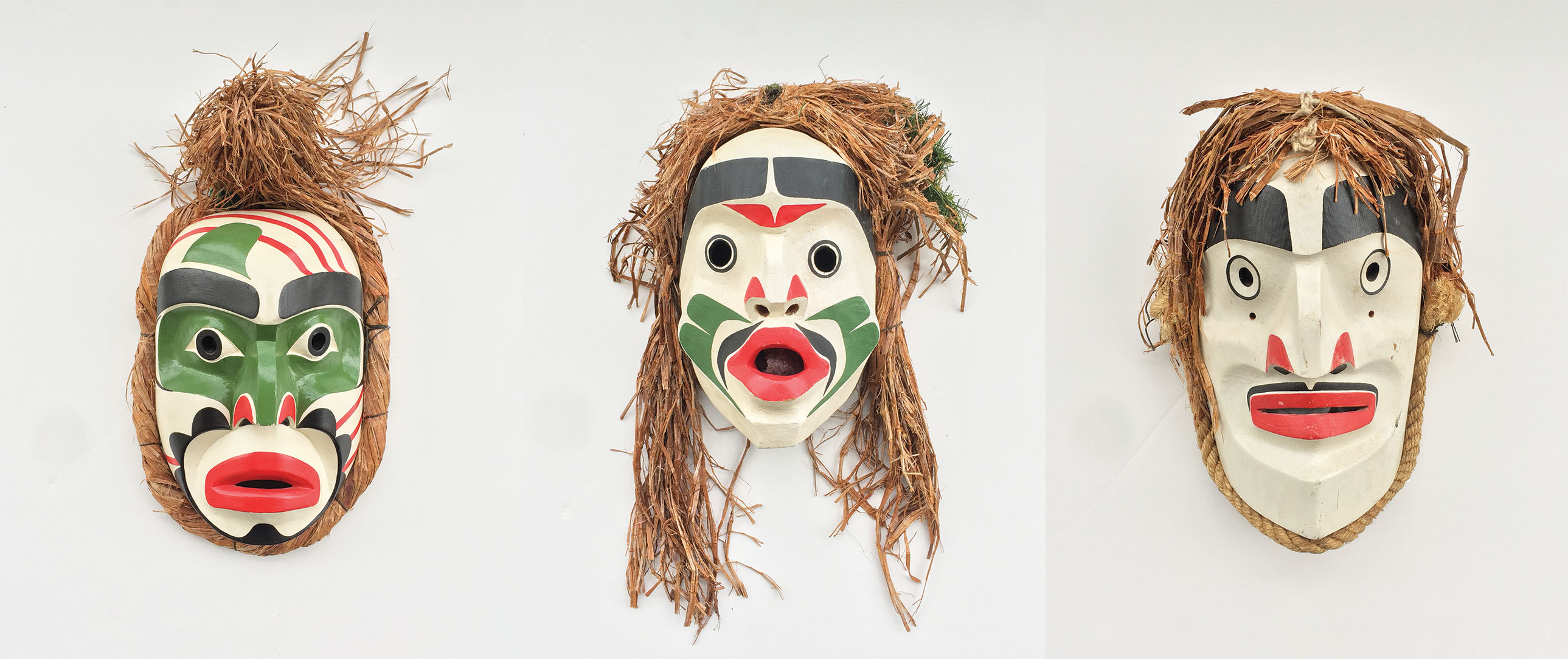 Three wooden masks from the same series with white, green, black, and red faces and straw-colored bark hair