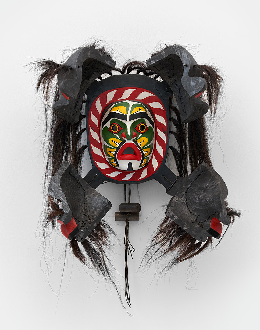 A wooden mask painted white, red, green, and yellow exposed under four split pieces of a cover