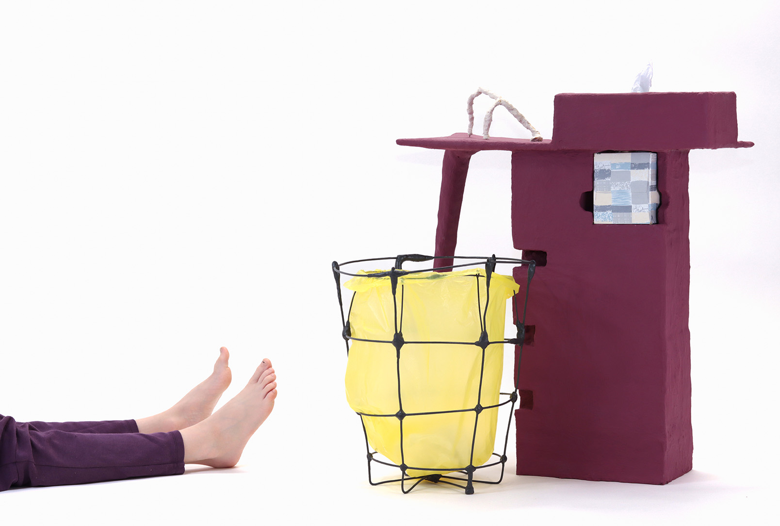 An artwork on the floor made up of a wire basket and a side table with a kleenex holder painted a deep maroonish purple. A small pair of legs and feet appear from the left of the image