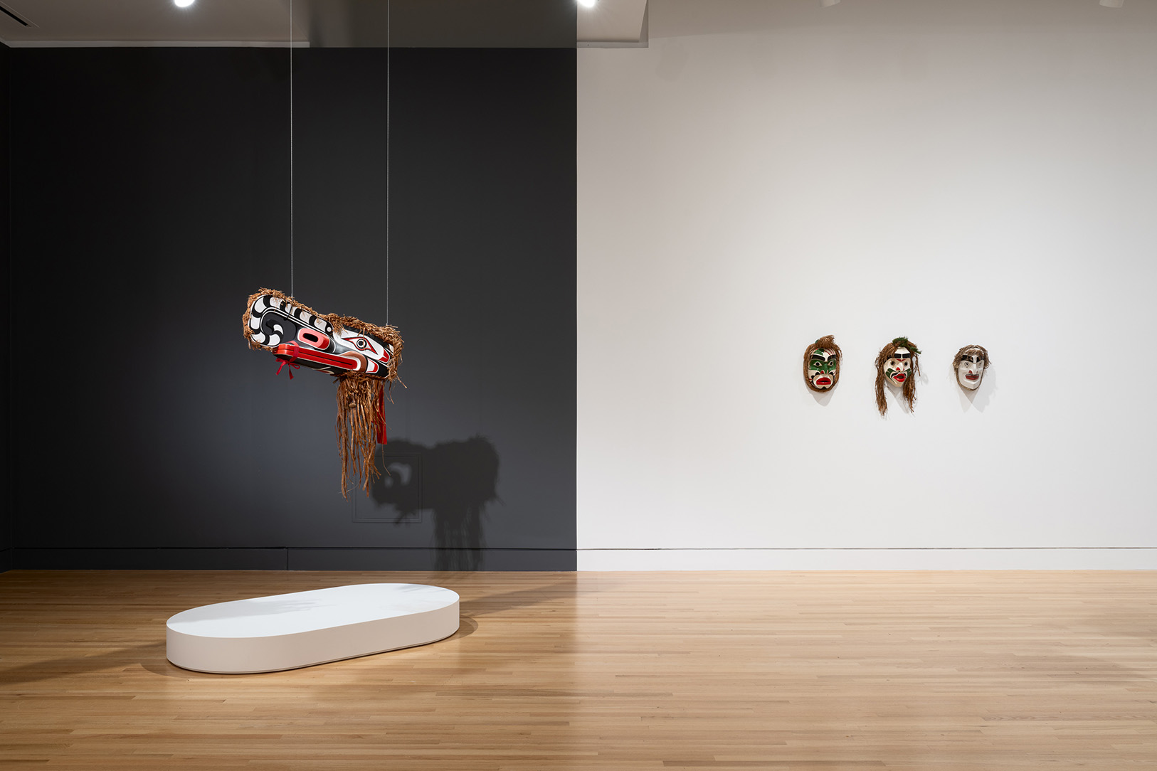 Artworks in a gallery space. On the left in front of a black wall is a hanging animal mask artwork, on the right are three smaller, humanoid masks