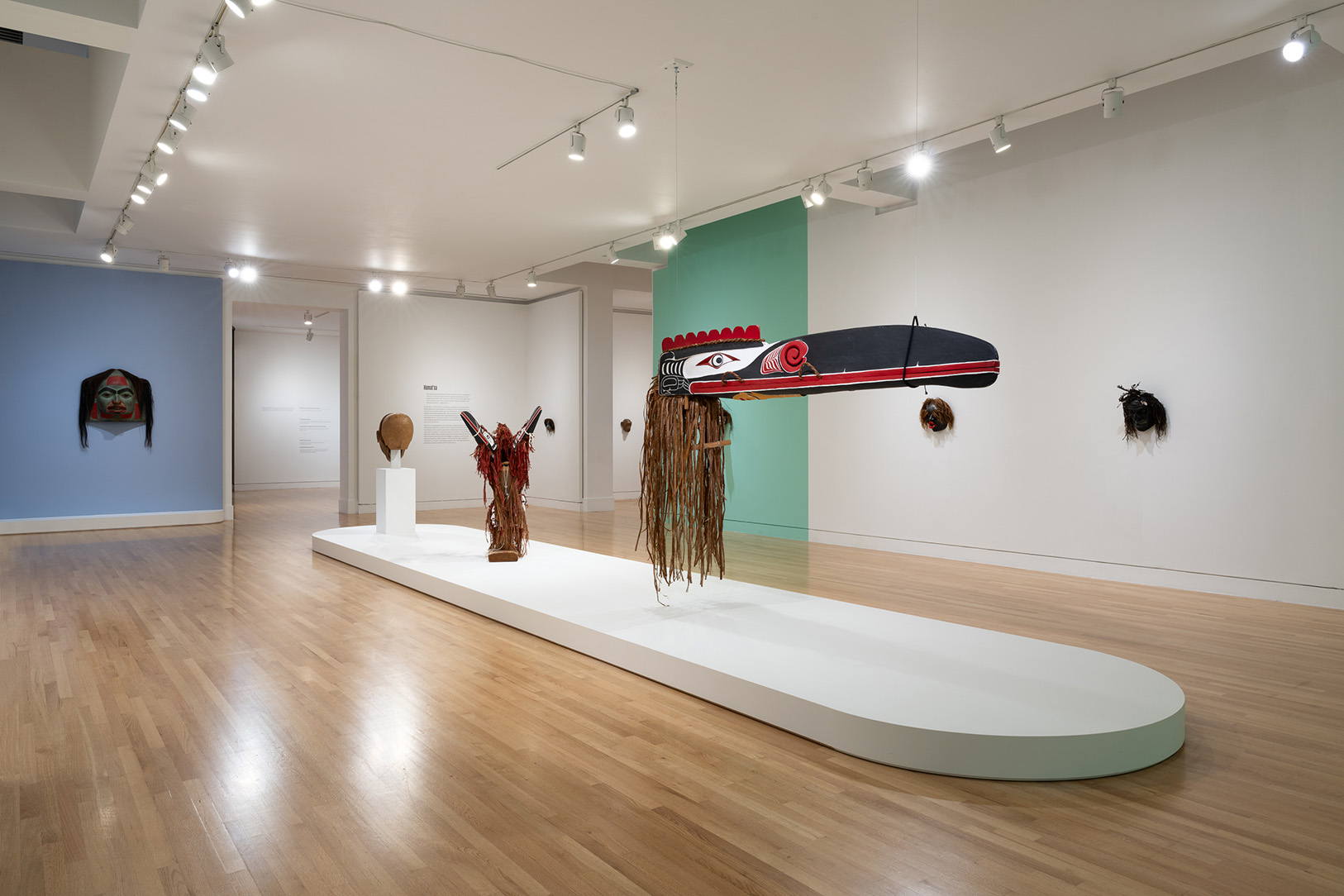 A gallery space with a number of wooden masks in the center and on the walls. Hanging from the ceiling in a prominent spot is a large, beaked animal shaped mask