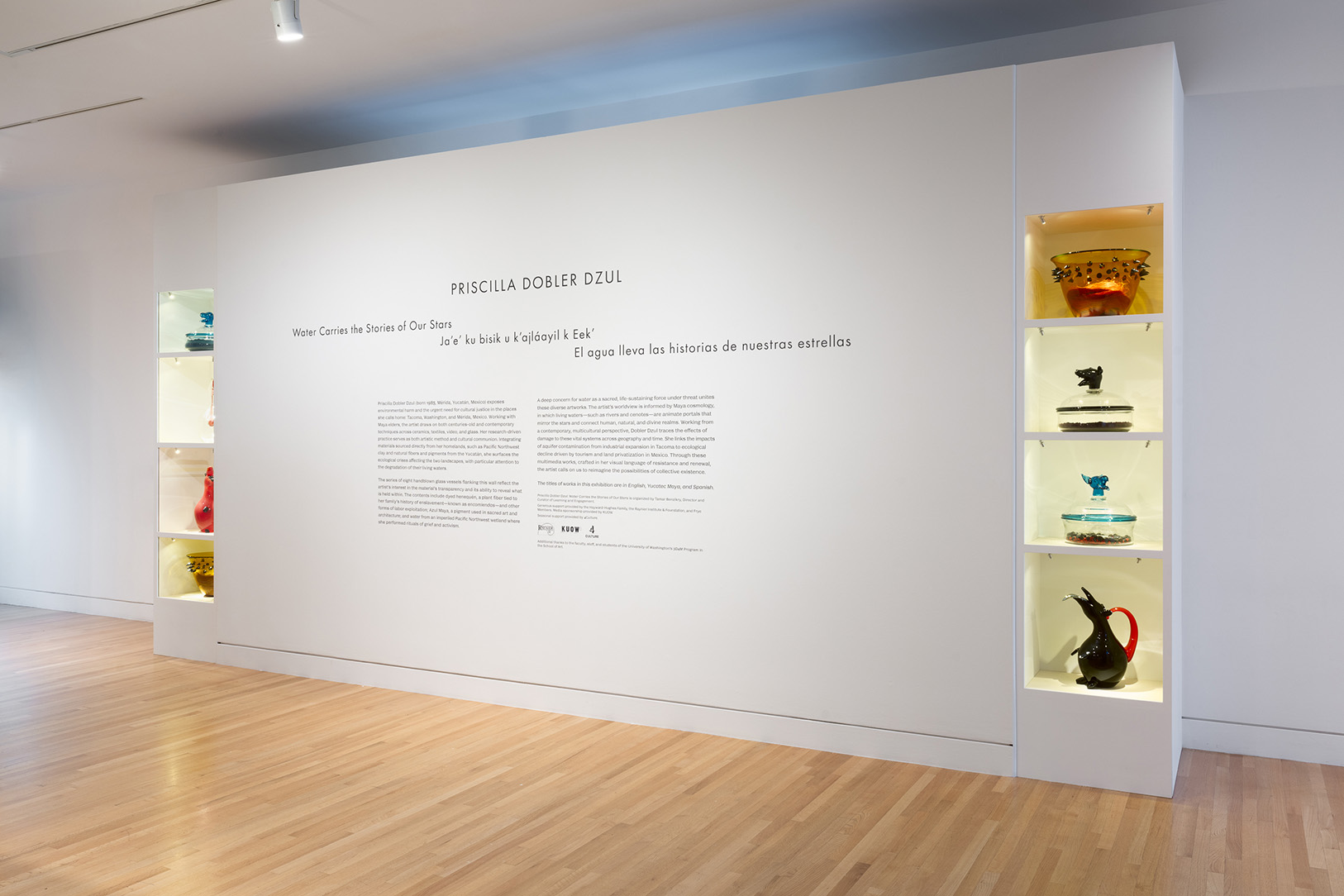 A white wall with text describing an art exhibition. On either side of the text are inset cabinets with colorful glass vessels