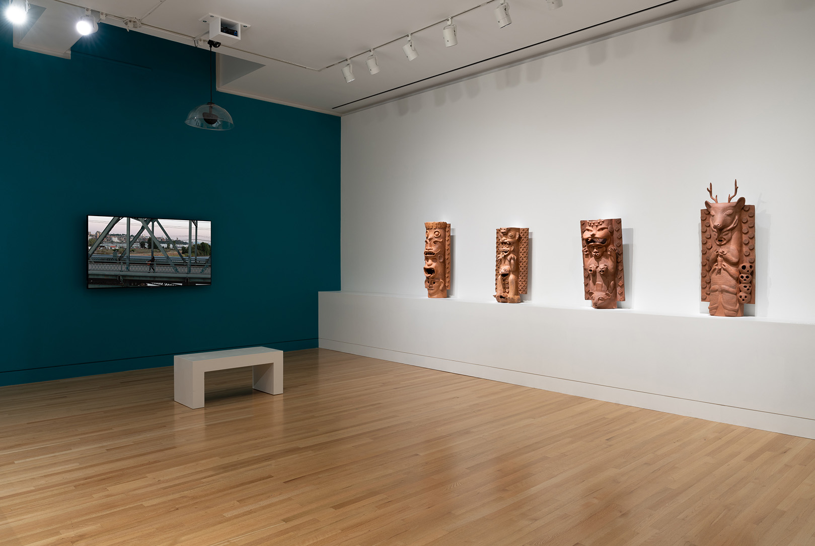 A corner of a gallery space. On one wall is a screen showing a video, and on the other is a pedestal with four large, freestanding ceramic sculptures
