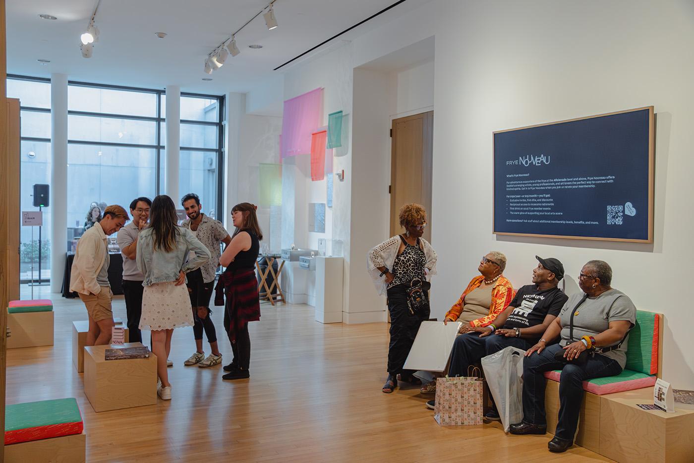 Groups of standing and seated people in a large parlor space with colorful benches and a large screen on the wall