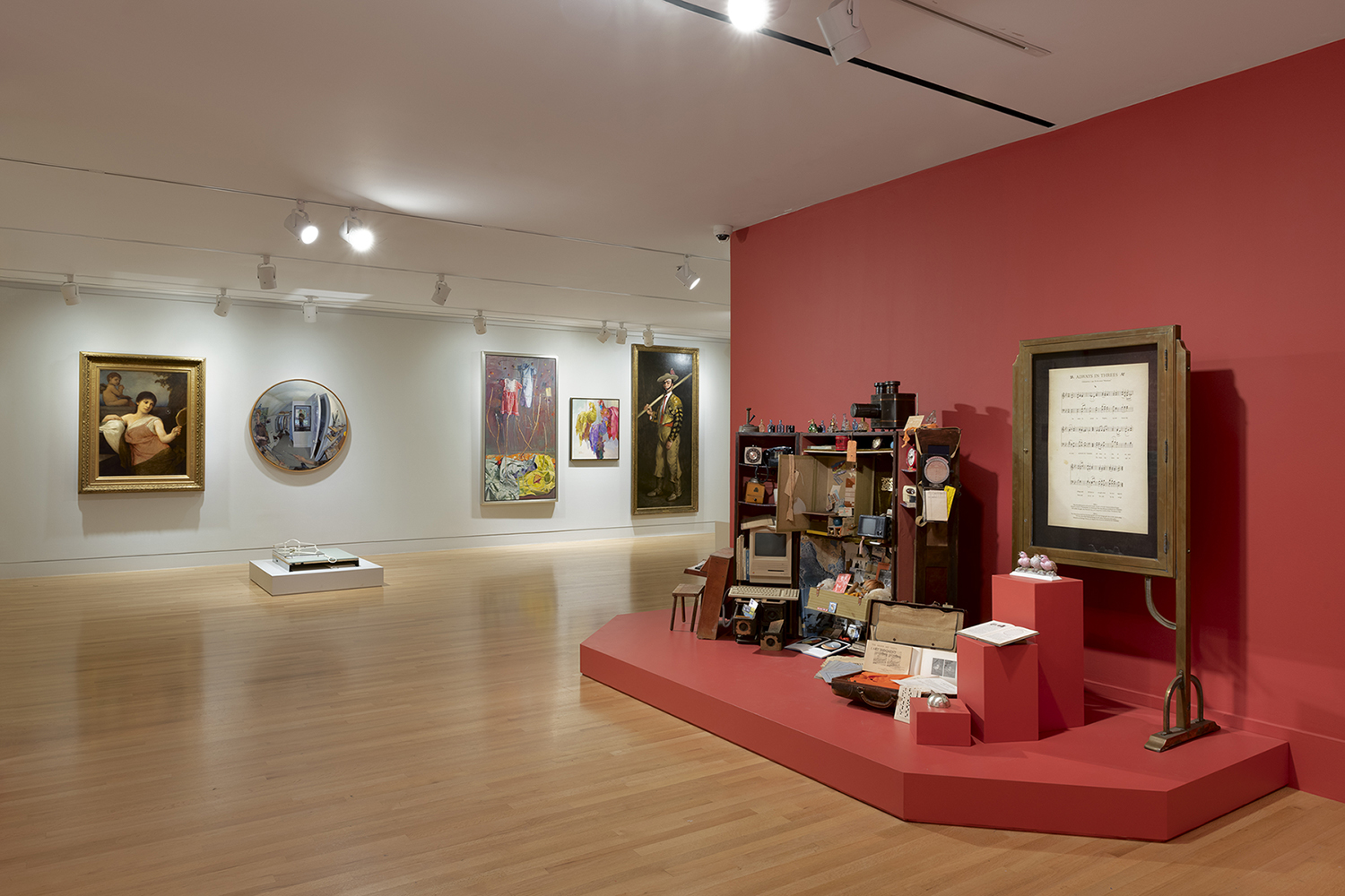 Several artworks hanging on a white gallery wall on the left, open trunk with miscellaneous items spilling out against a red wall on the right 