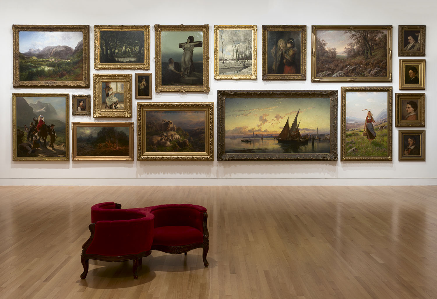 Paintings hung floor to ceiling in a gallery with white walls, one red gossip chair in the foreground