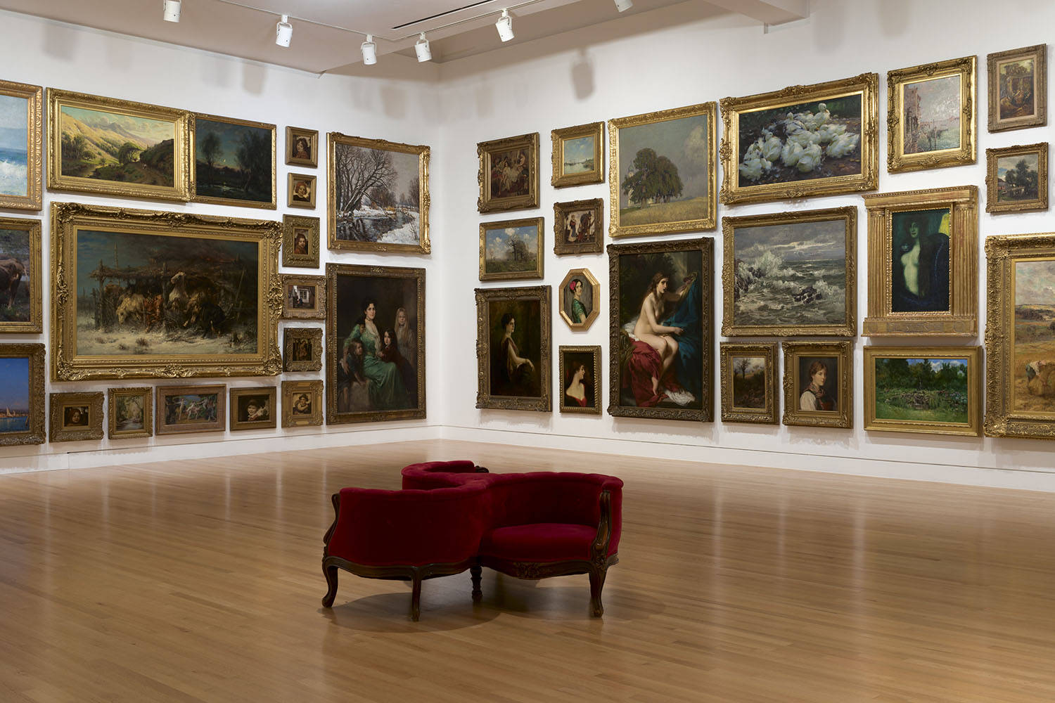 Paintings hung floor to ceiling in a gallery with white walls, one red gossip chair in the foreground