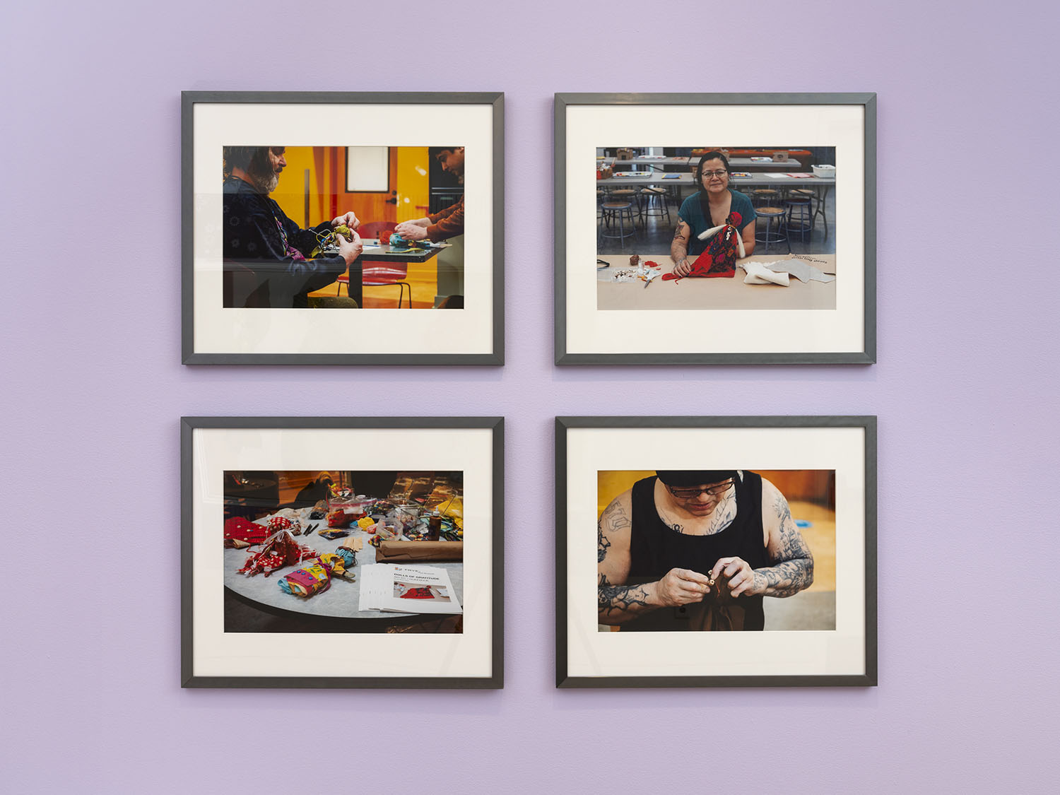 Four framed photos of people making dolls, installed on a light purple wall