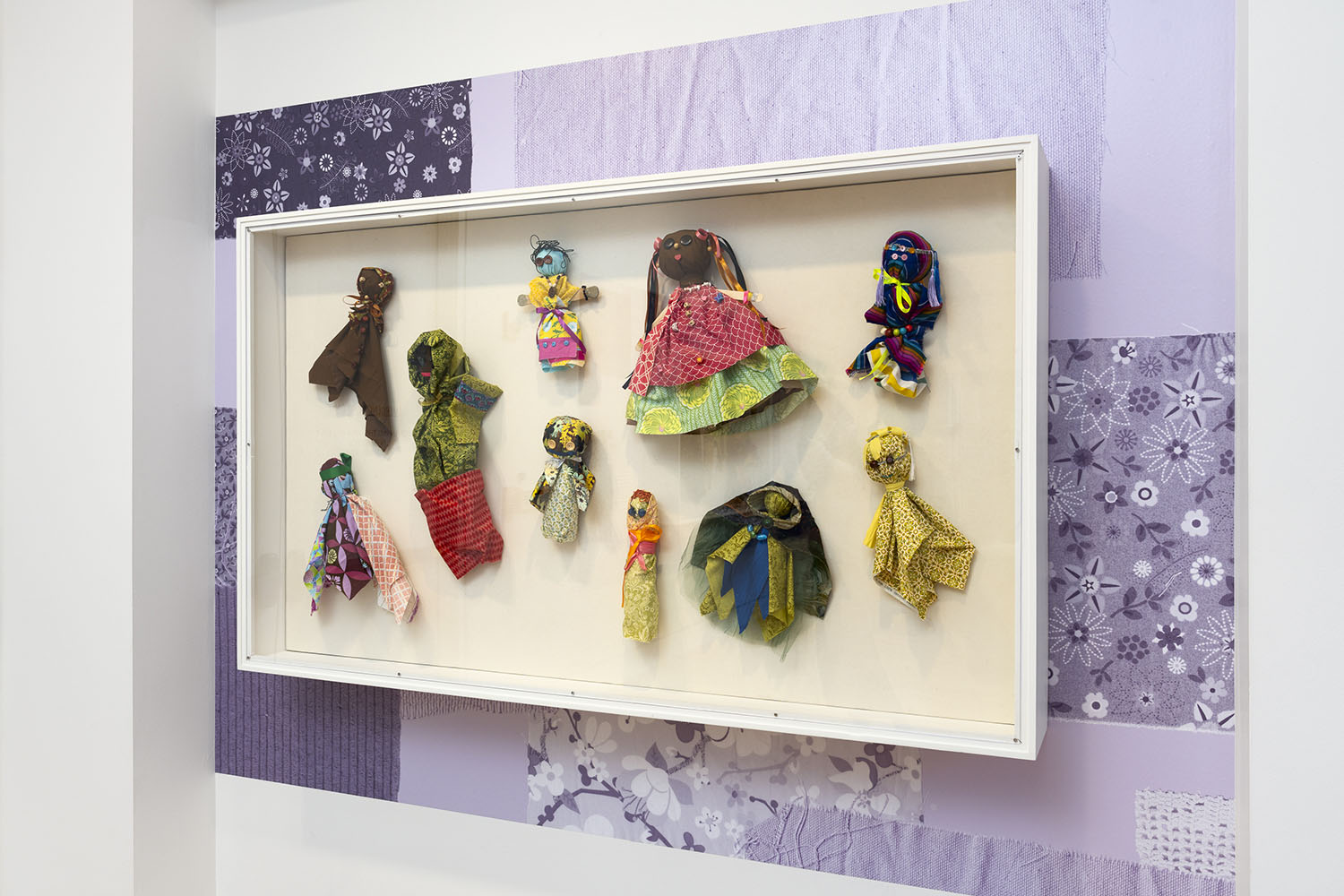 Photo of handmade dolls in a glass case, installed on top of a purple super graphic with images of fabric pieces