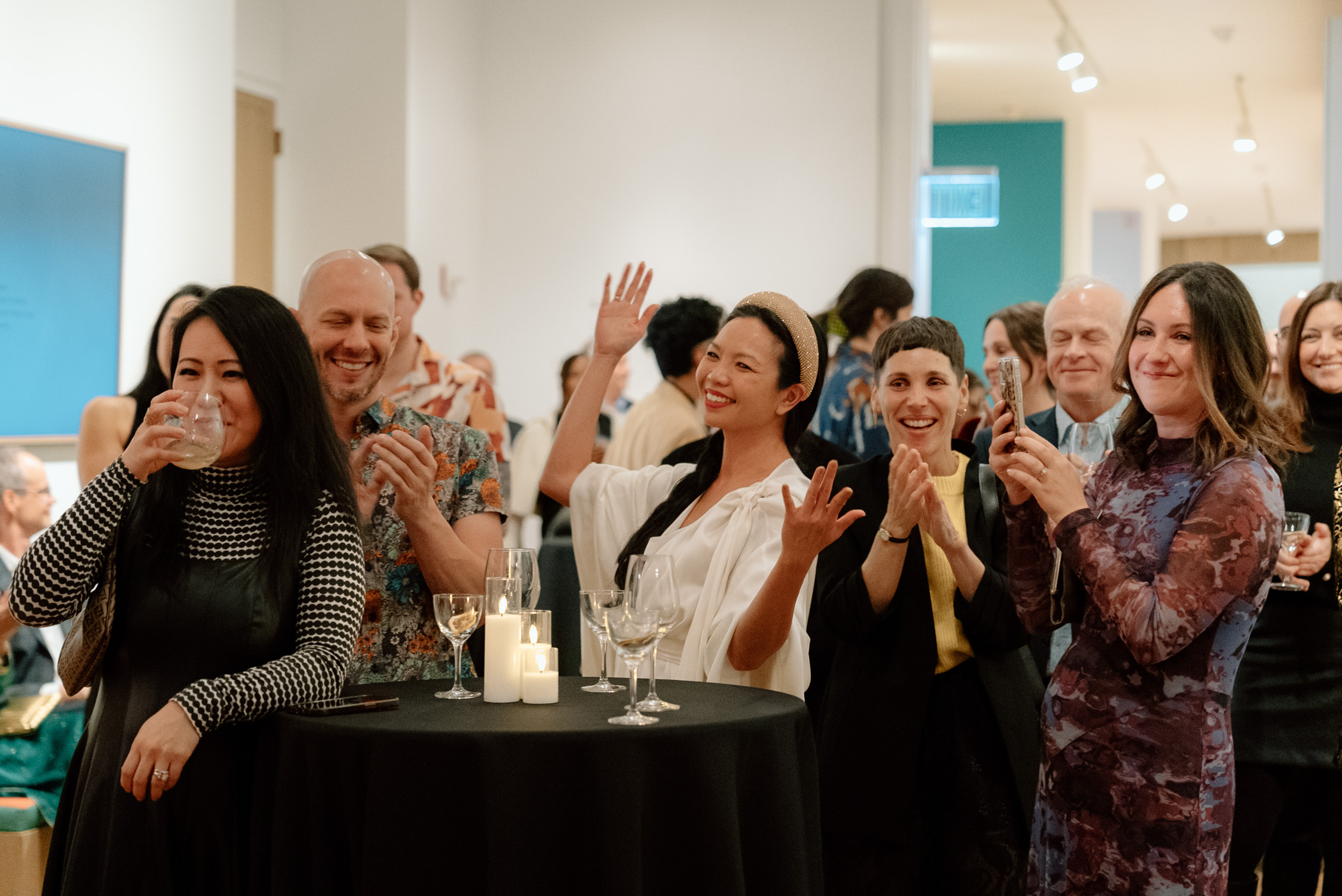 A large group of people standing by a tall table and smiling while listening to someone out of the frame