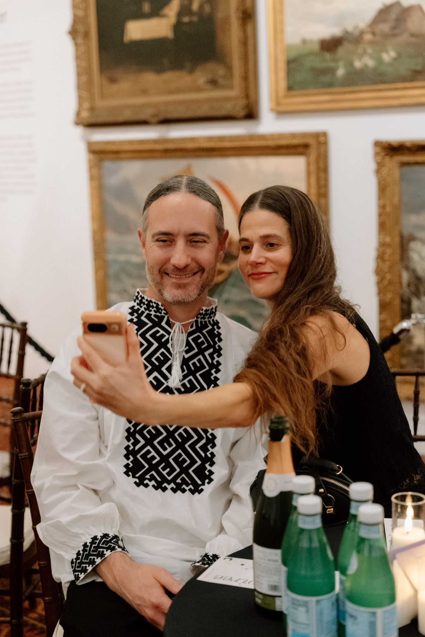 Two people sitting and smiling in front of oil paintings hanging on a wall