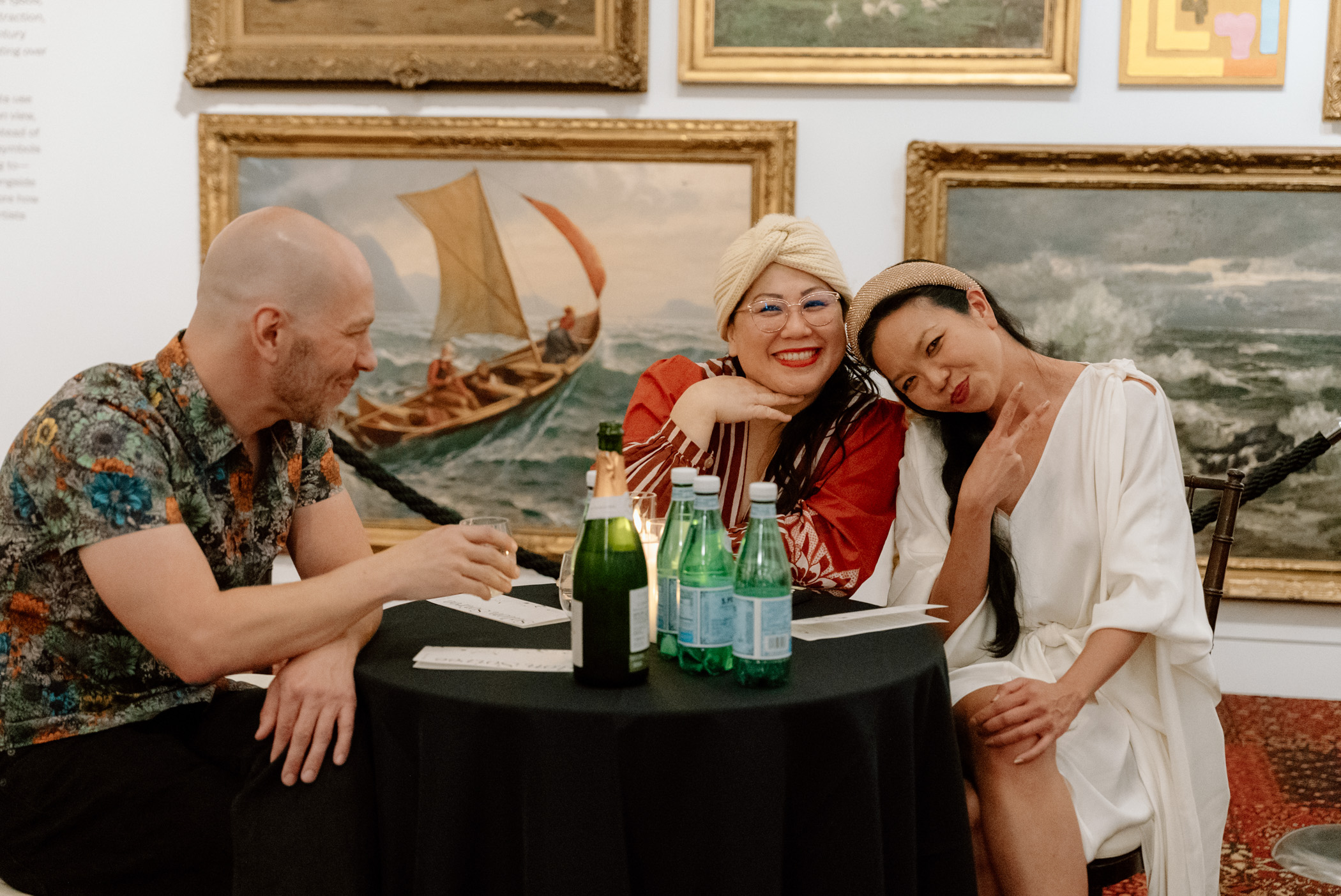 Three people sitting and smiling in front of oil paintings hanging on a wall