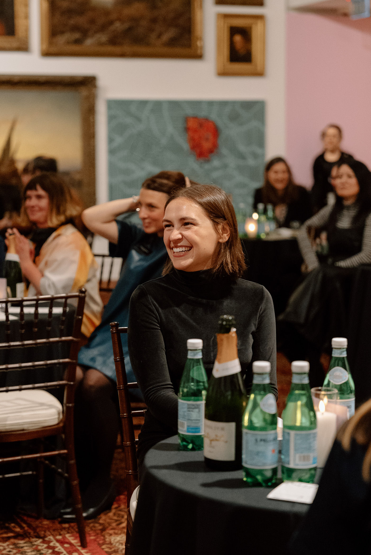 A young woman seated at a table and smiling in a room whose walls are covered in paintings