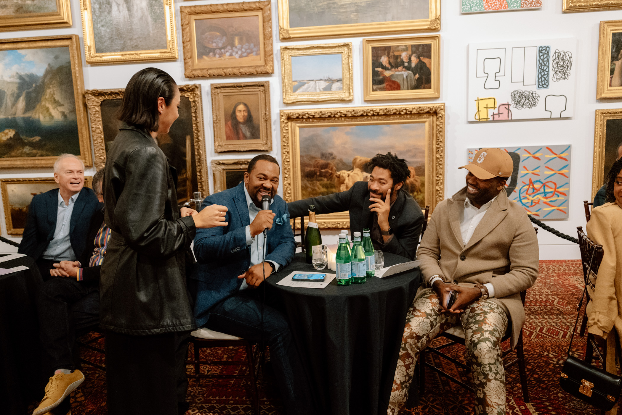 People sitting at a table in front of a wall covered in paintings. One is holding a microphone and speaking.