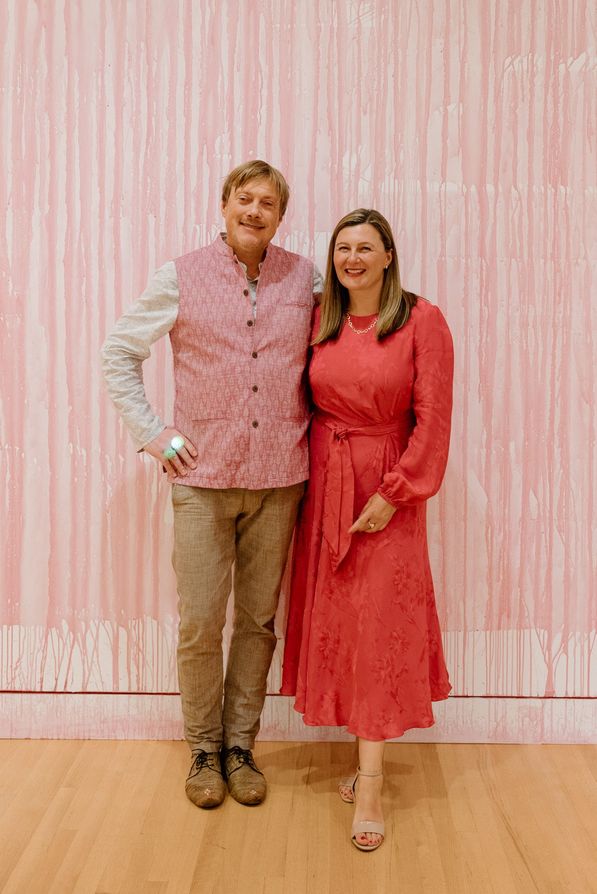 A couple smiling and posing in front of a wall covered in dripping pink paint