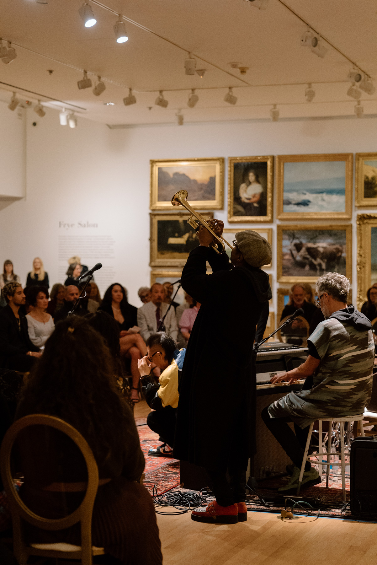 A person photographed from behind raising a trumpet that he's playing in front of a wall covered in paintings with opulent gold frames