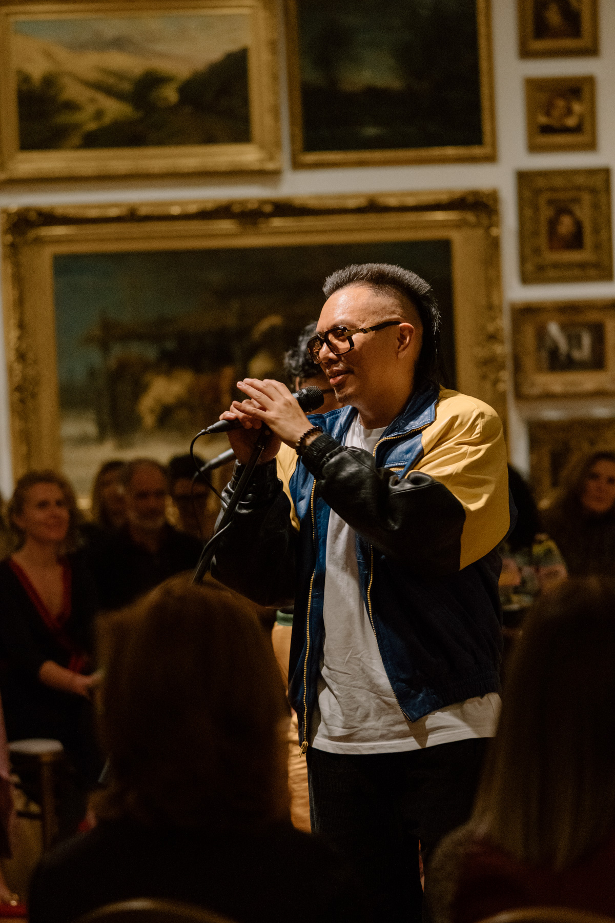 A person performing with a mic in front of a wall covered in paintings with opulent gold frames
