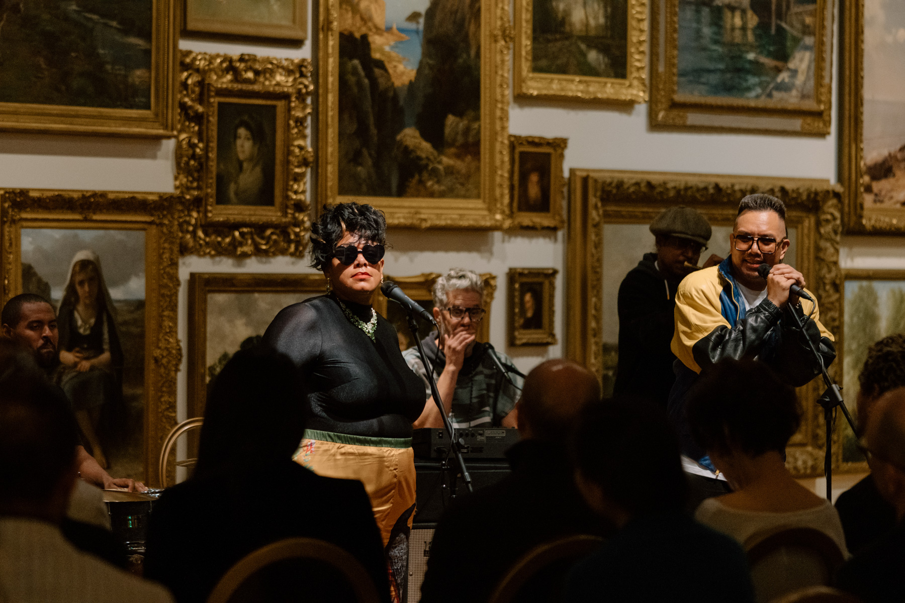 People playing music and signing in front of a wall covered in paintings with opulent gold frames