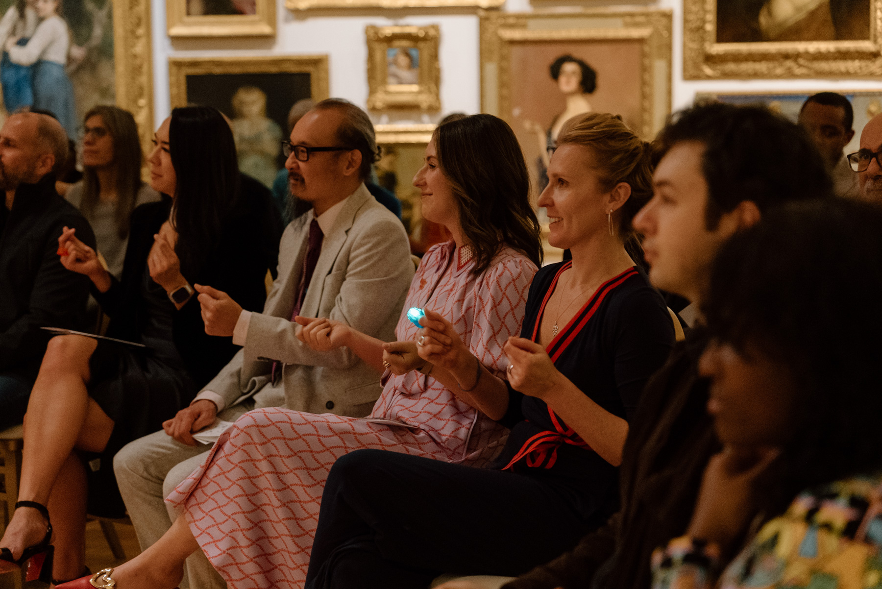 People sitting and watching a performance in front of a wall covered in paintings with opulent gold frames