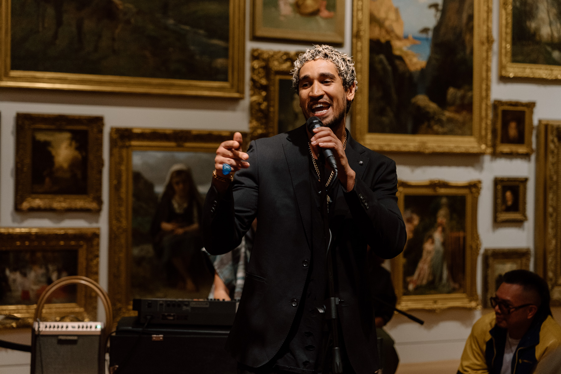 A person performing with a mic in front of a wall covered in paintings with opulent gold frames