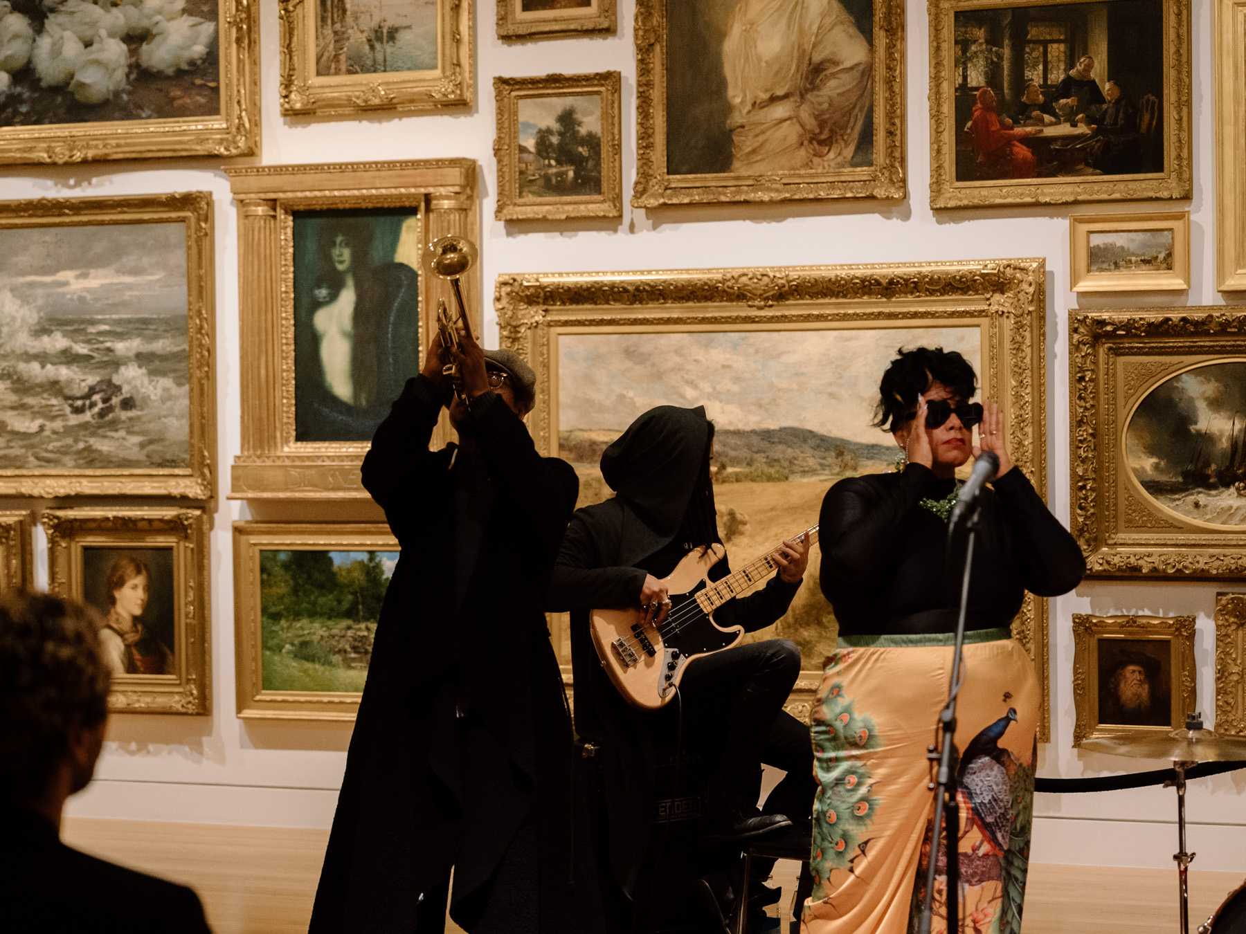 People playing music in front of a wall covered in paintings with opulent gold frames. One is playing a trumpet, another is drumming, and a third is signing