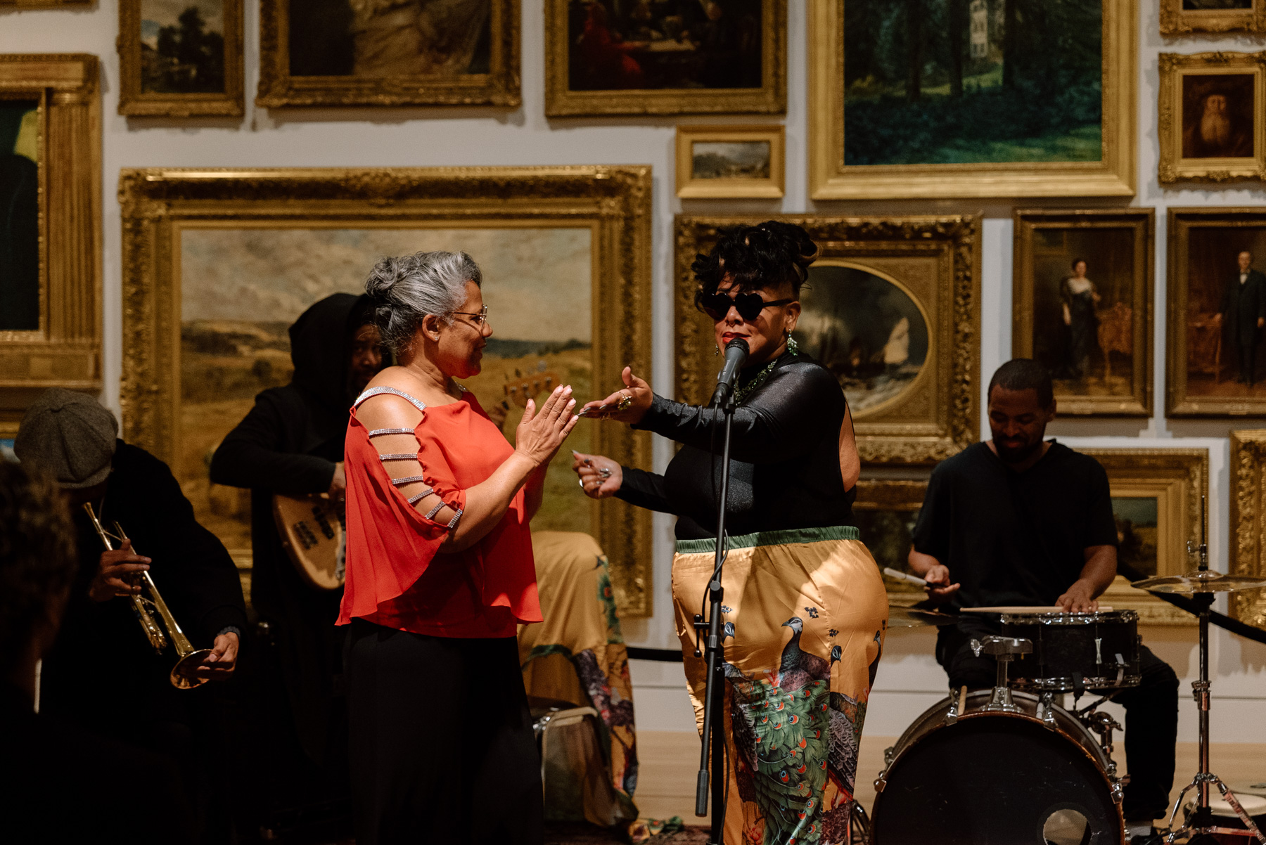 People performing in front of a wall covered in paintings with opulent gold frames