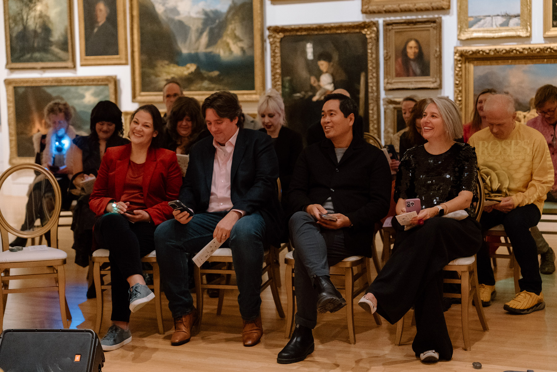 People sitting in chairs and watching a performance in front of a wall covered in paintings with opulent gold frames
