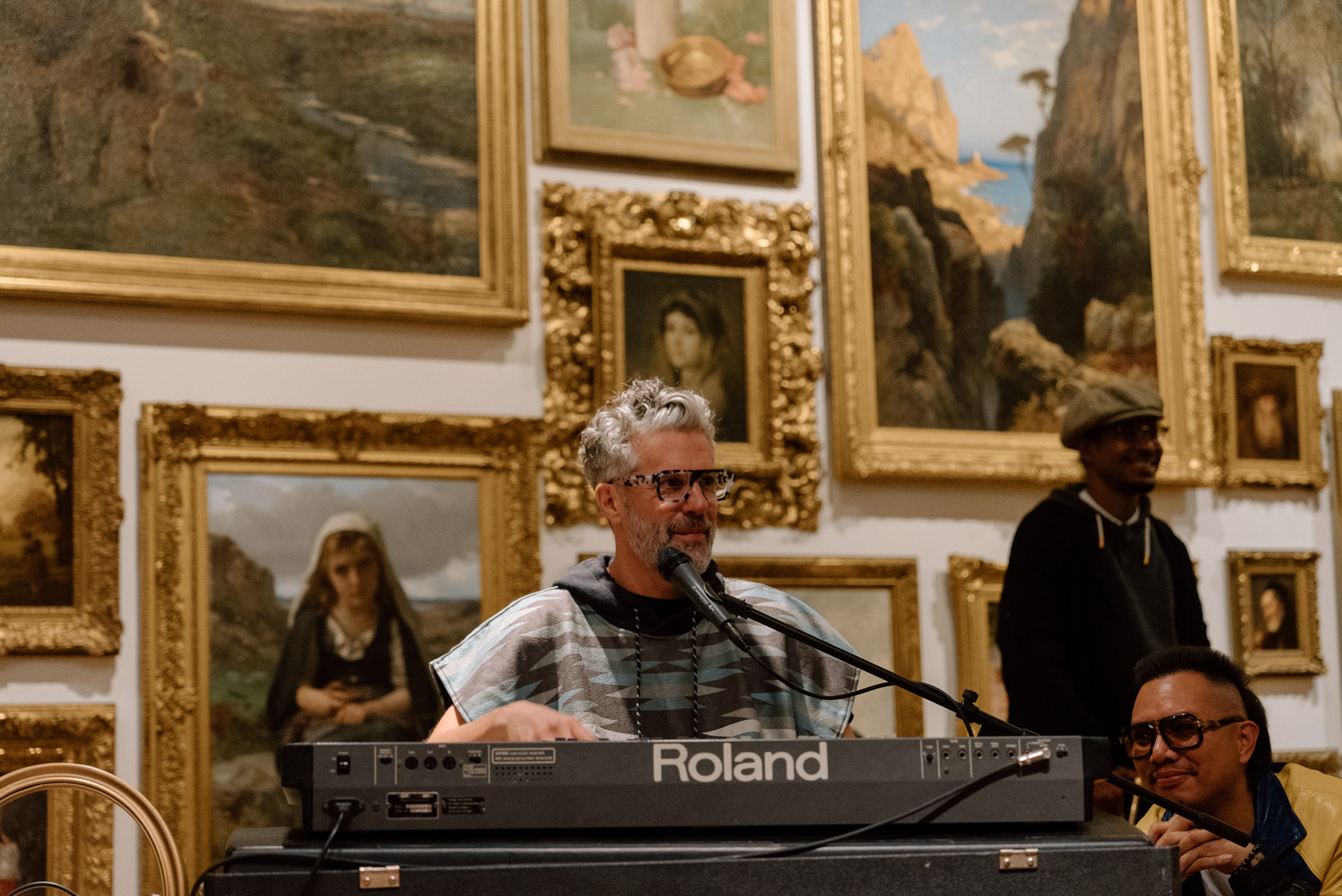 A person playing a keyboard and signing into a mic in front of a wall covered in paintings with opulent gold frames