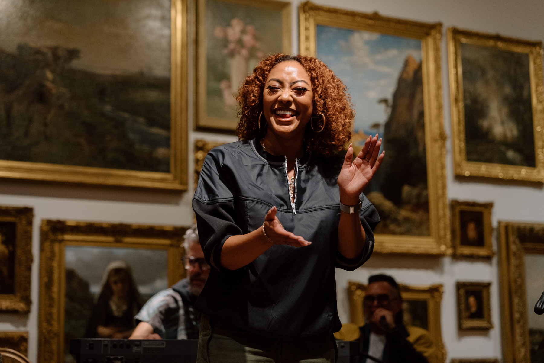 A person smiling and clapping in front of a wall covered in paintings with gold frames