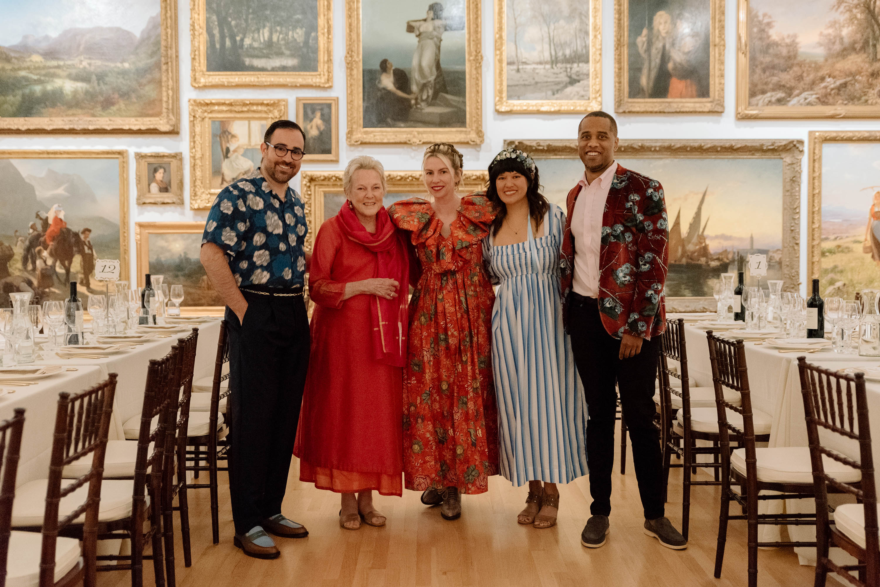 A group of people posing for a photo between tables in a large room with paintings on the walls 