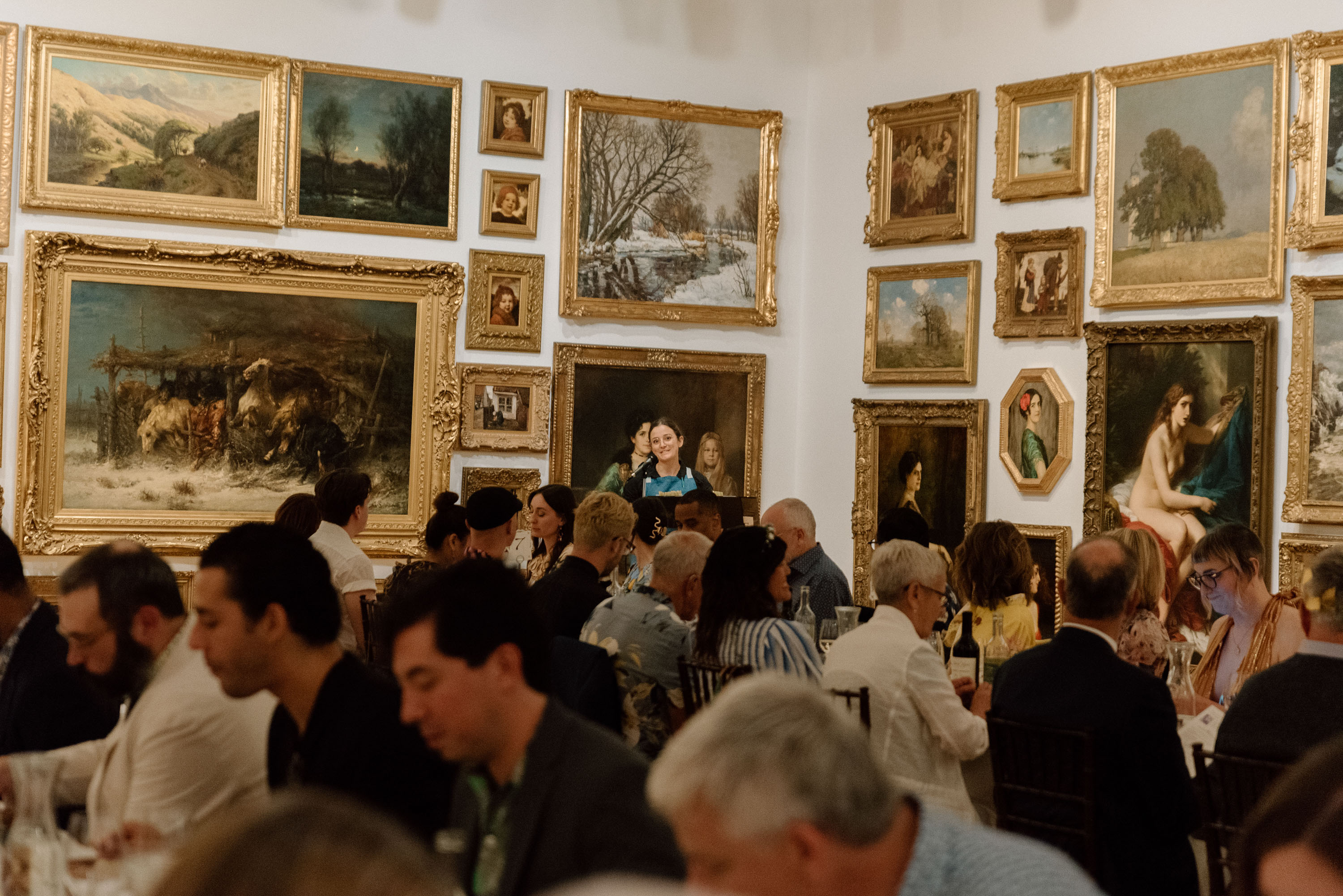A person standing in the background of a busy room with paintings on the walls and people sitting at tables in the foreground