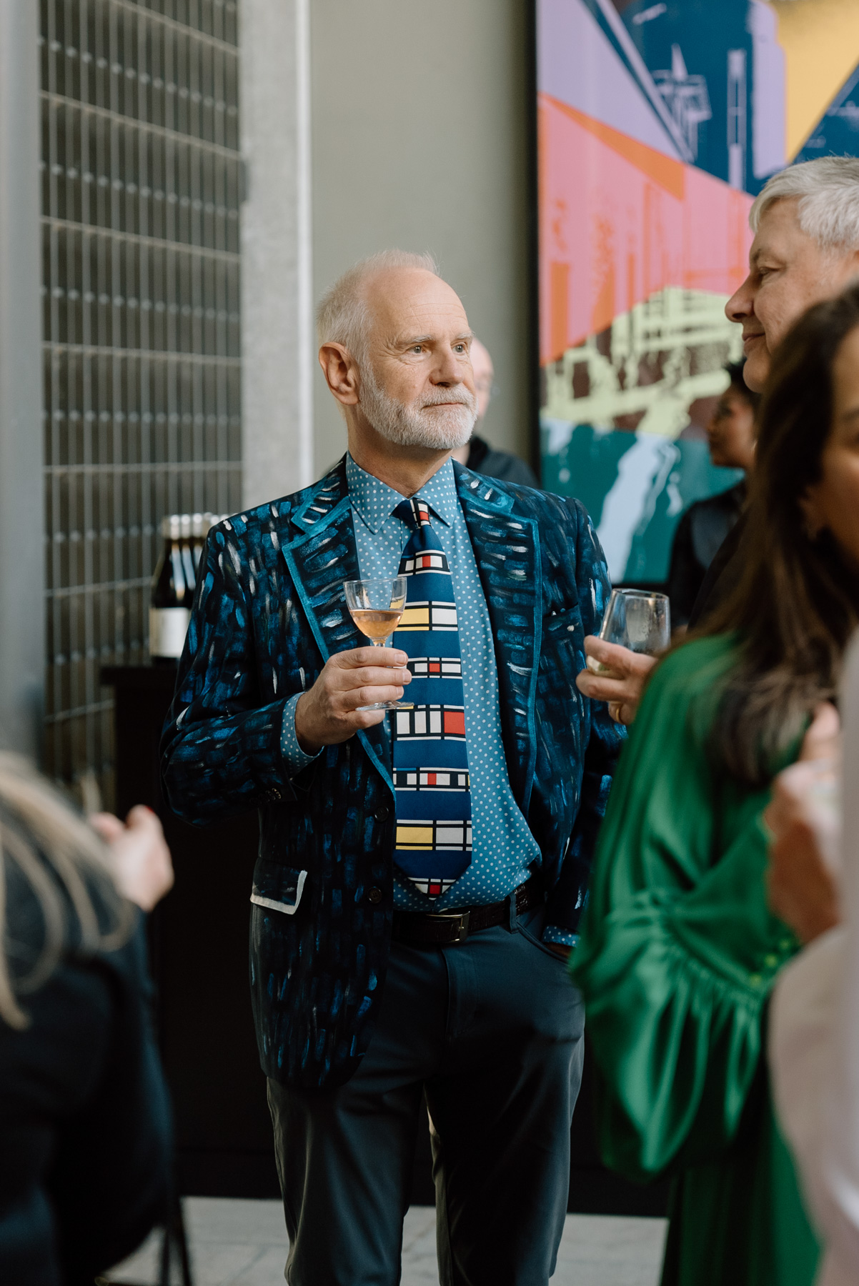 An older man wearing a colorful suit jacket and tie holding a glass of wine and listening to another person