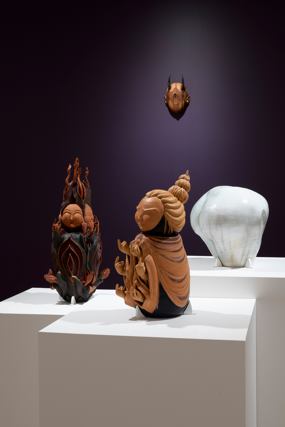 Three ceramic vessels on while pedestals and a ceramic mask hanging on a purple wall behind them