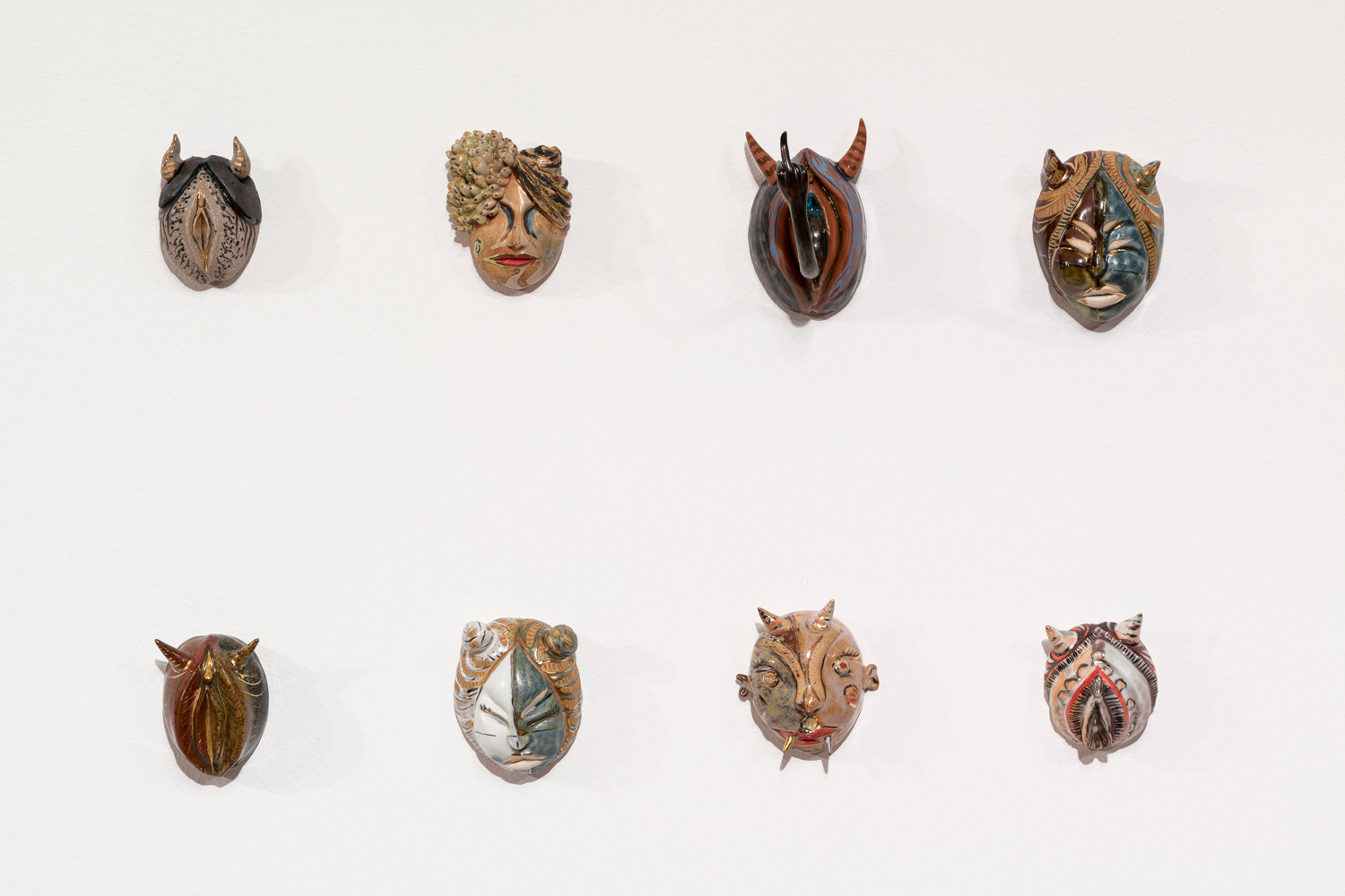 Eight small ceramic masks in two rows with various motifs including vulvar shapes and demonic horns on a white wall