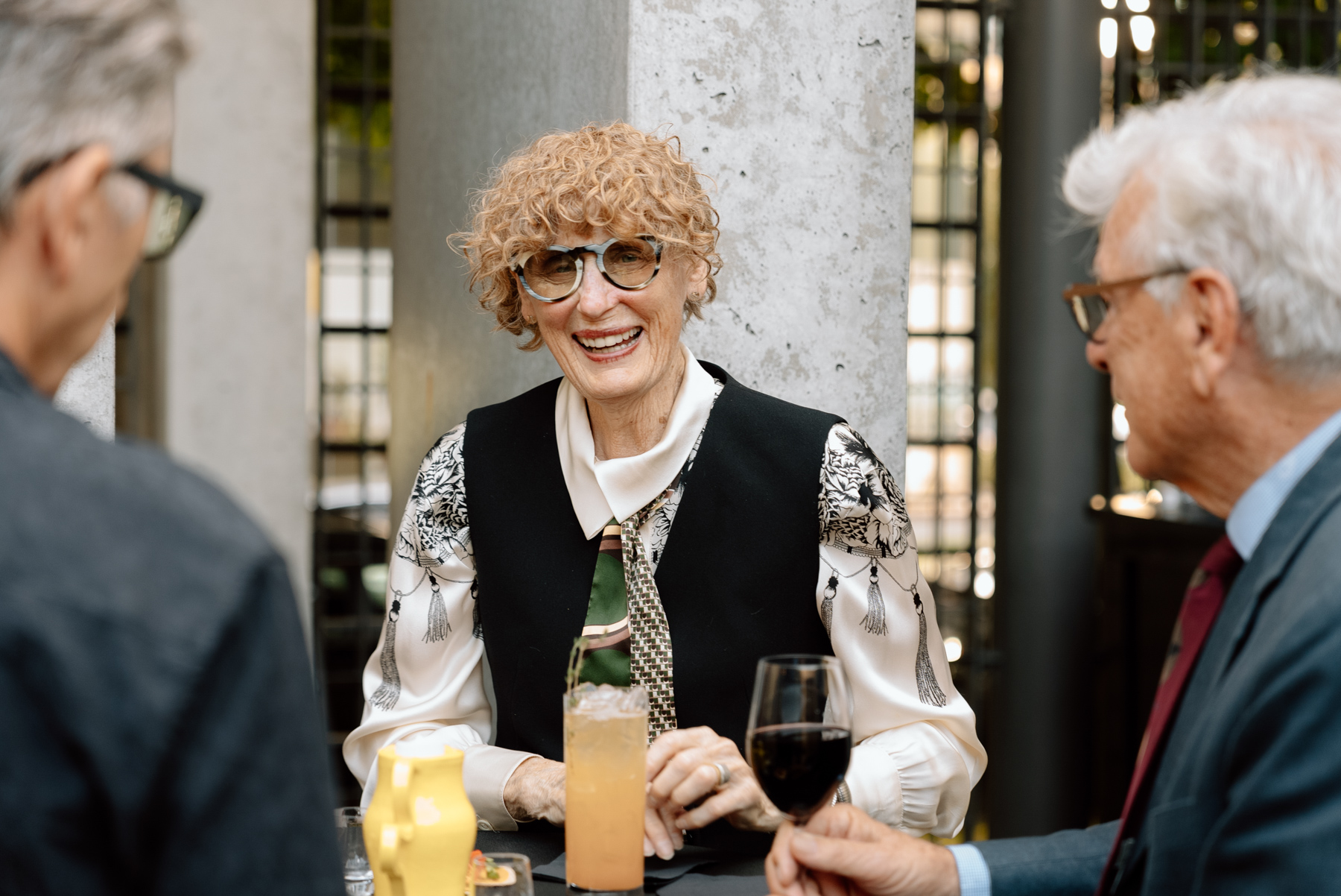 Three people in cocktail party attire standing around a small table with drinks and chatting and smiling