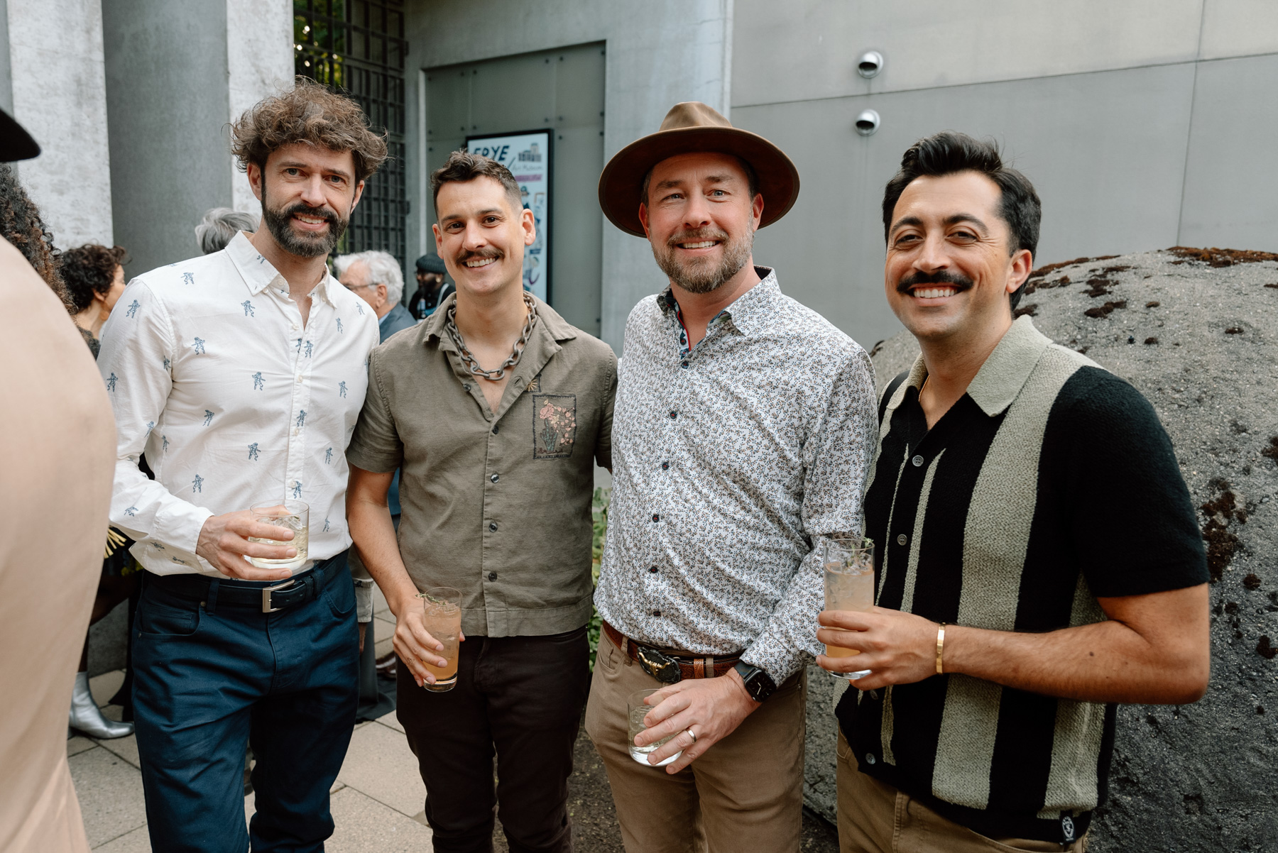 A group of four men in cocktail party attire smiling and posing with drinks in a courtyard area