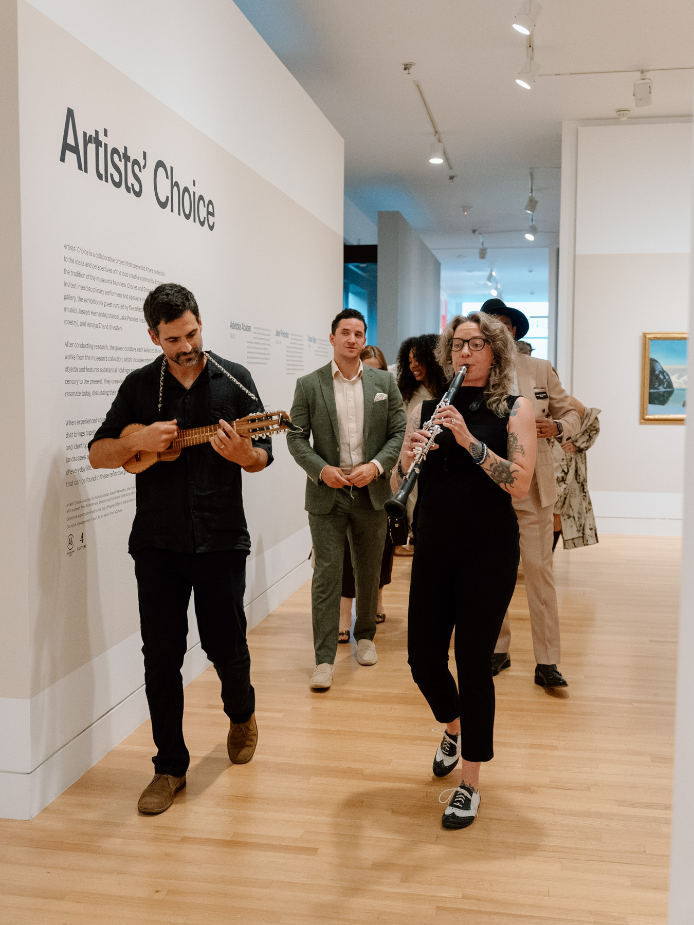 A pair of musicians playing a ukelele and a wind instrument leading a group through an art museum
