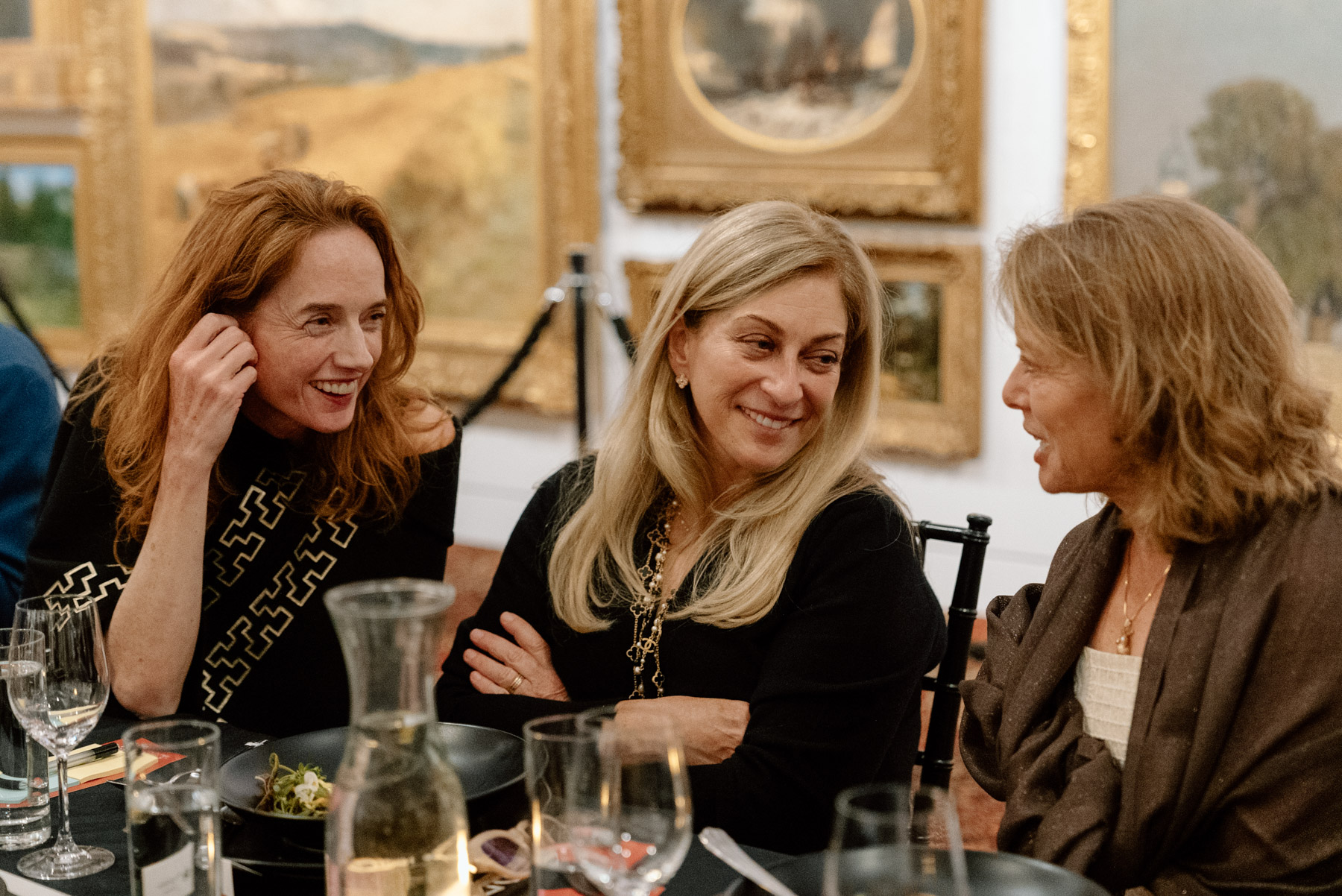A group of seated women smiling and chatting at a long table in a gallery space