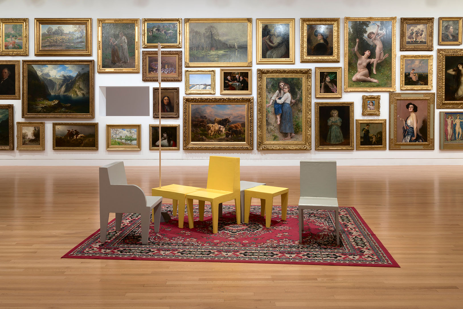 A group of yellow and gray seats in front of a wall covered in paintings. A rectangular hole is cut in the wall.