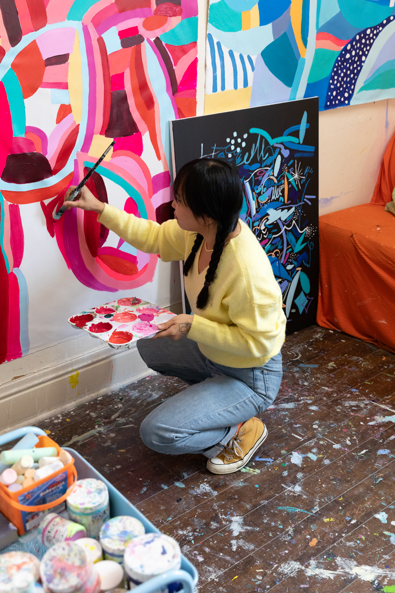 Photo of Nikita Ares, a woman with dark brown hair, painting with bright pink paint while crouching