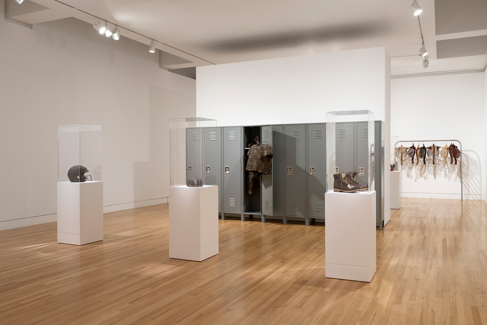 An art gallery room with a set of high school lockers behind a series of artworks of clothing items covered in layers of tree bark