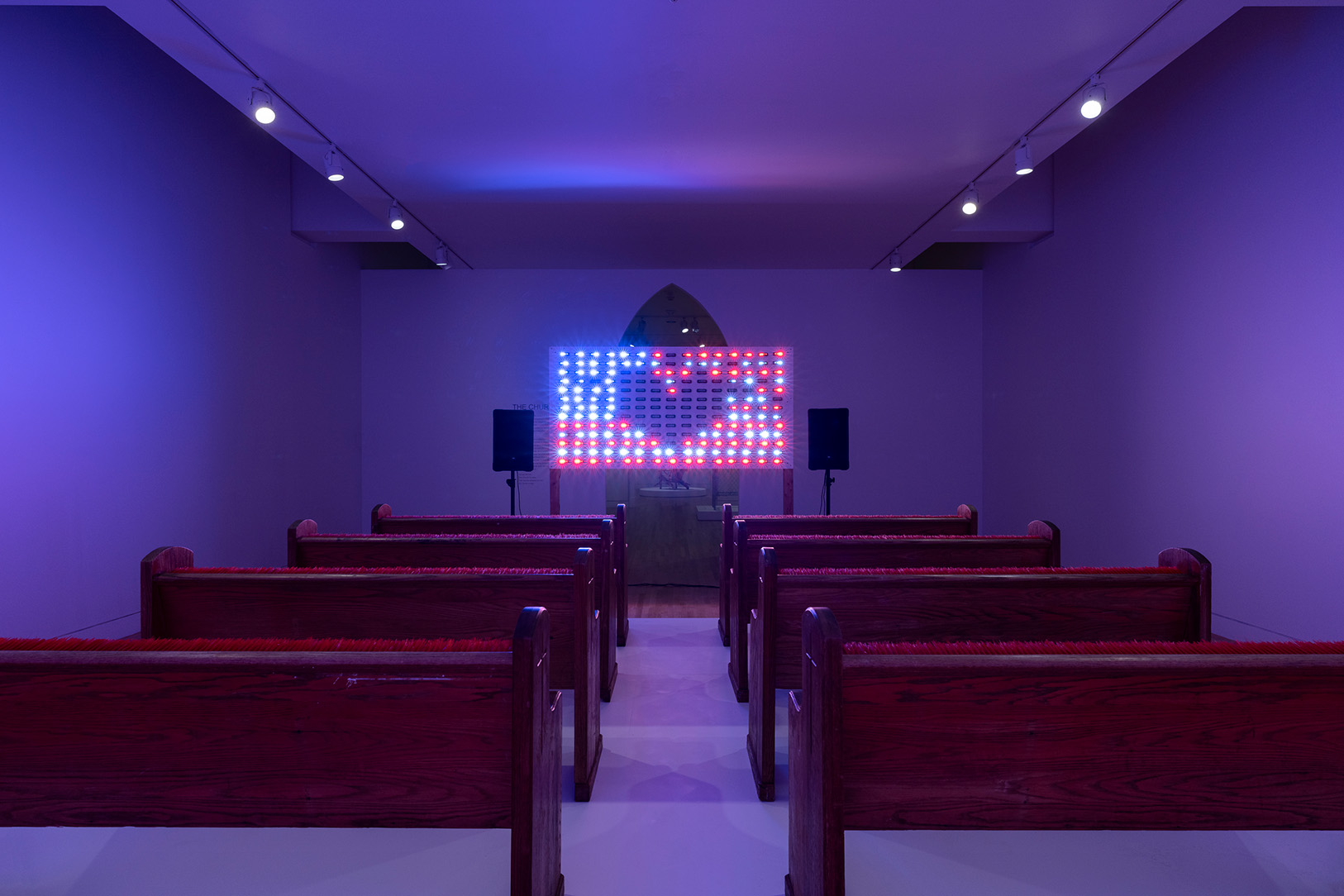 A dark room in an art gallery with church pews facing a screen of individual lightbulbs in the general shape of an American flag